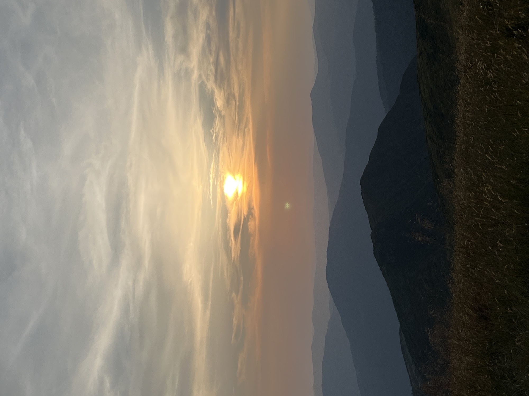 The image depicts a mountainous landscape at sunset. The sky is filled with clouds, with the sun low on the horizon, casting a warm, orange glow. Layers of mountains recede into the distance, their silhouettes becoming darker and bluer. In the foreground, tall grasses are visible, suggesting an outdoor, natural setting. The scene is calm and serene, characteristic of a remote, natural environment. While the provided hints are helpful for focusing on specific details such as the sky and foreground, the overall image captures a vast expanse of nature. The description provided does not include specific details about Luhy, Ukraine as the image itself does not provide such information.