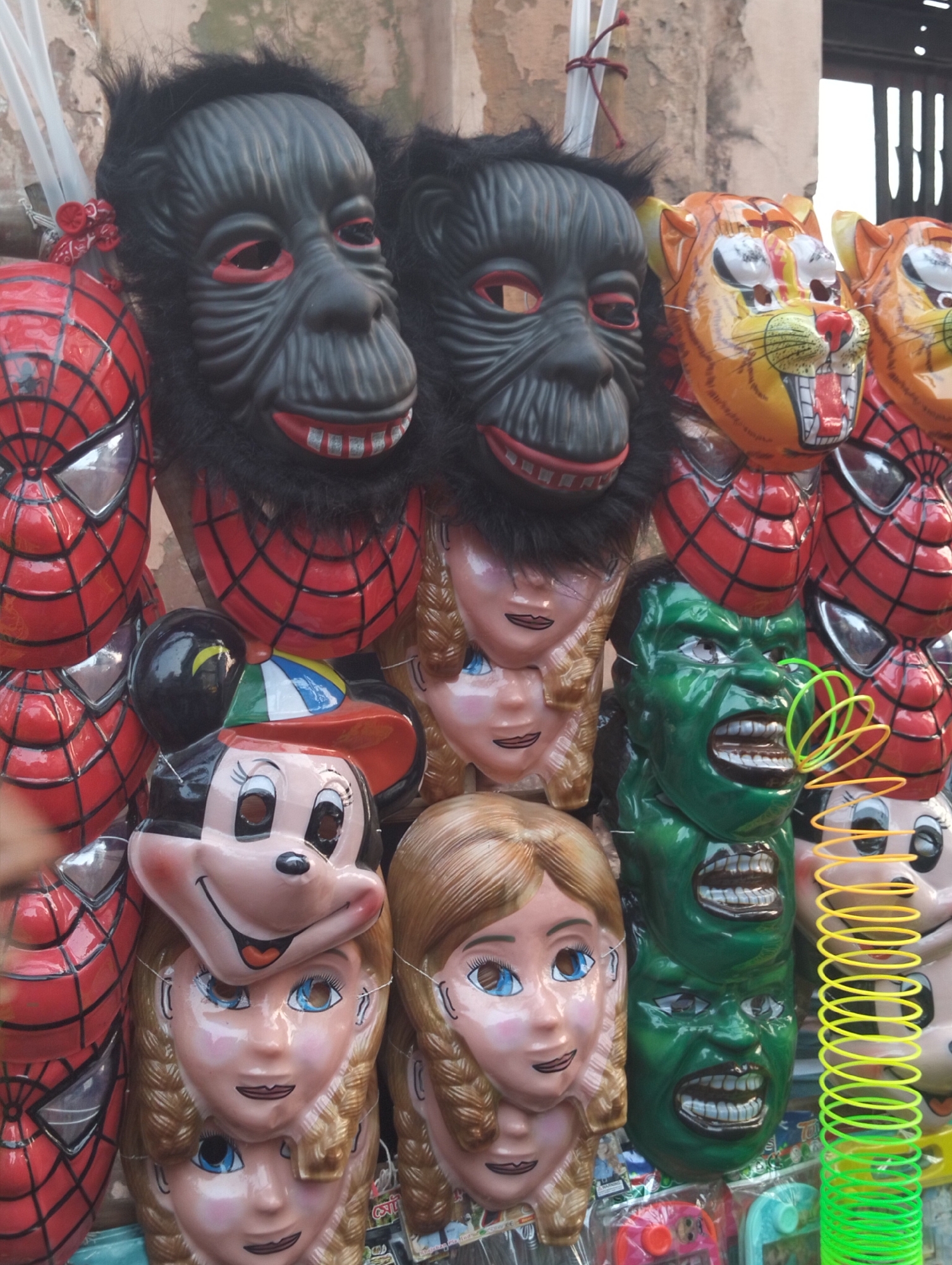 A still shot shows a display of character masks for sale in what is likely a street market in Dhaka, Bangladesh. Masks of popular characters such as Spiderman, The Hulk, a tiger, a gorilla, Minnie Mouse, and a generic princess character are arranged vertically against a wall. A plastic slinky toy is near the edge of the display. The masks appear to be made of molded plastic with painted details, and some have faux fur elements (gorilla). The scene suggests an outdoor market setting.