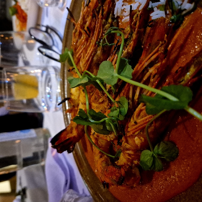 A close-up shows a plate of cooked prawns in a reddish sauce, garnished with green sprigs. Rice grains are scattered among the prawns. A table setting is visible in the background with glassware containing a yellow liquid, and a pair of glasses. The prawns are arranged closely together, with their reddish-orange color dominating the dish.