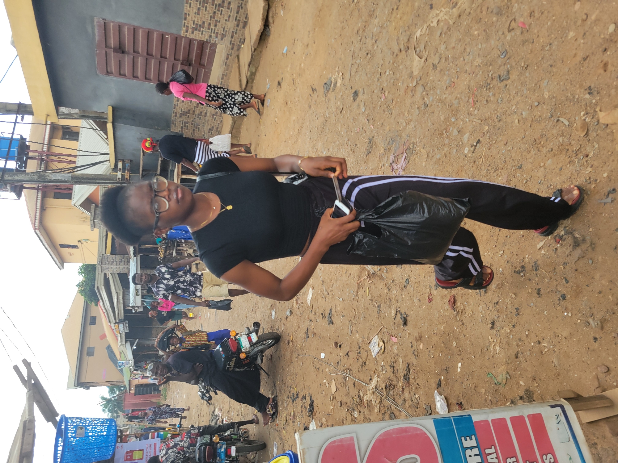 The image captures a street scene in Egbagu, Nigeria, likely during the daytime with overcast skies. The foreground features a woman wearing glasses, a black t-shirt, and black athletic pants with white stripes. She is holding a black plastic bag and a smartphone. Her posture suggests she might be walking or paused.
The background reveals a bustling market or street with several other individuals and various objects. To the left, a blue plastic basket is visible. Further back, there are buildings with corrugated iron roofs and some are painted yellow and grey. A few motorcycles are parked, with one in the mid-ground bearing multiple items strapped to it.
Other people in the scene include a woman in a pink top and a patterned skirt walking away from the camera, and another person wearing a black and white striped shirt. A man in a black and white patterned shirt is also visible in the mid-ground. The ground is dusty and appears to be a dirt road with scattered debris.
The overall impression is of a vibrant, everyday scene in a Nigerian community, possibly a market day or a busy period for local commerce. The lighting suggests it is either early morning or late afternoon. There is no clear indication of specific emotions, but the general atmosphere seems active and ordinary. A partial view of a sign with red and blue lettering is visible at the bottom right, but the text is too fragmented to read.