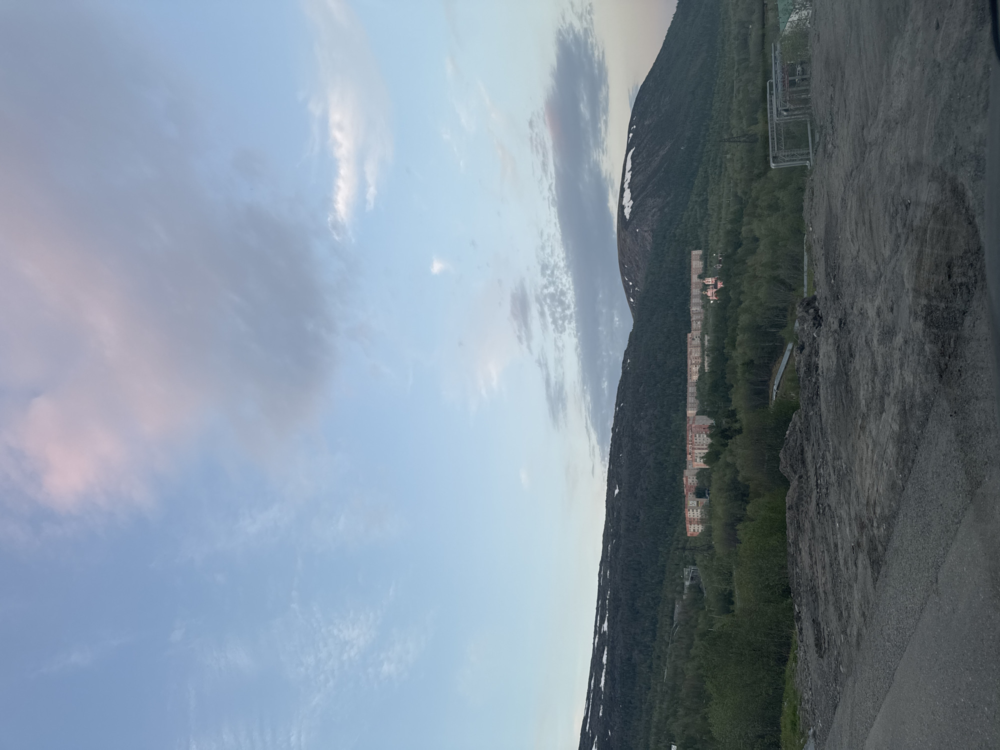 The image depicts a panoramic view of a landscape identified as Kirovsk, Russia, under an overcast sky with elements of sunrise or sunset light. The sky occupies the upper half, displaying varying shades of light blue with scattered stratocumulus and altocumulus clouds. A large, diffuse cloud mass with pink and purple hues is visible in the upper right quadrant.
The middle ground is dominated by a range of hills or low mountains. The slopes are covered predominantly by dense, dark green coniferous forest. Patches of snow are visible on the upper slopes and peaks of the mountains, particularly on the rightmost peak.
Nestled within the forested foothills are several urban structures. A cluster of multi-story residential buildings, likely apartment blocks, with light-colored facades (beige, off-white, orange-pink) and red roofs, are visible towards the center-left. To the right of these residential buildings, further back in the forest, stands a distinct pink or red building with multiple domes, suggesting a religious structure such as a church or cathedral, though its specific function is not definitively observable. Industrial-like structures or pipelines are also visible among the trees in the lower-right midground.
The immediate foreground consists of rough, disturbed earth and gravel, appearing as an undeveloped or construction site. A dark asphalt road surface is partially visible along the bottom-left edge. No human figures or active vehicles are present in the scene. The overall impression is one of a semi-urbanized area set against a natural, mountainous, subarctic or boreal environment.