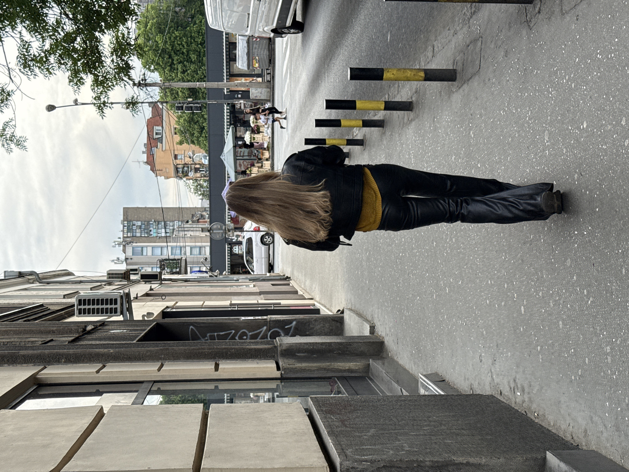 A woman with long blonde hair, wearing a black jacket and black pants, walks on a street in Vracar Urban Municipality, Serbia. The street has black and yellow bollards and a storefront to the left. In the background are buildings, a white van, and people walking towards what appears to be a marketplace or vendor area.