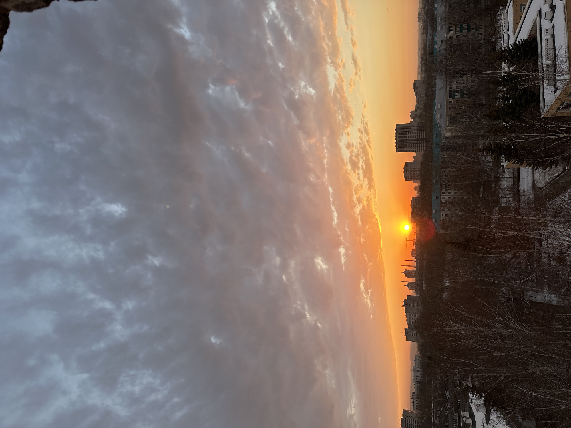 The image captures a sunset in what appears to be Kazan, Russia. A line of buildings, including residential and industrial structures, sits against a bright orange horizon with the sun visible between them. Bare trees in the foreground and along the roofs of buildings are partially covered with snow, suggesting a winter setting. The sky above is filled with layers of clouds, transitioning from shades of grey to orange nearer the sun. The overall scene gives a sense of urban setting in the cold season.