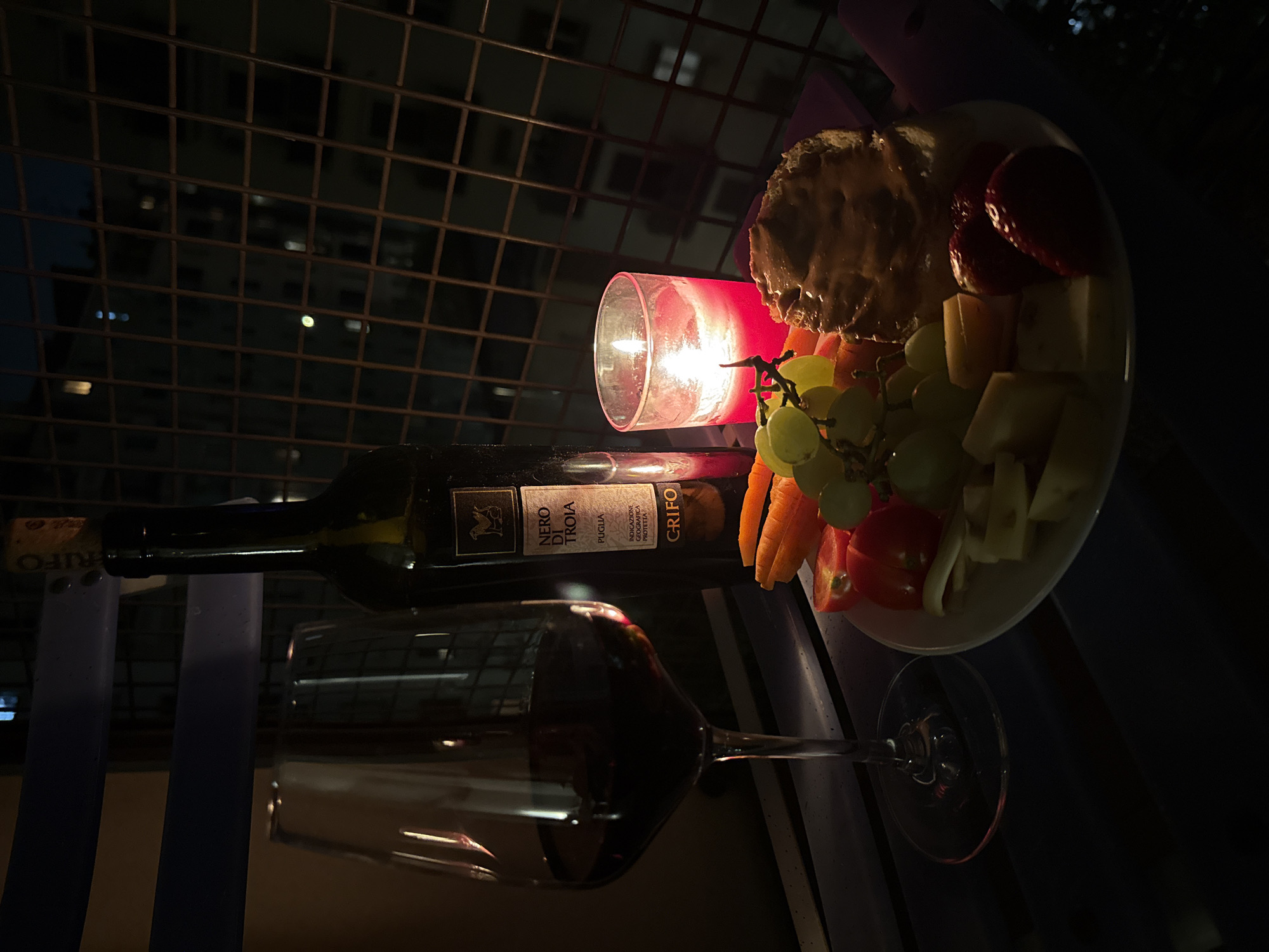 Here's a description of the image:
**Scene:** An intimate outdoor setting, possibly a balcony, at night. A wine bottle labeled "Nero Di Troia," a wine glass filled with red wine, a lit candle in a red glass holder, and a plate of snacks (cheese, crackers with topping, strawberries, grapes, carrots, tomato slices) are arranged on a surface. Behind the scene is a dark building with a metal grid in front.
**Location:** The presence of "Nero Di Troia" wine from Puglia, Italy suggests the location is possibly in Italy. However, without further identifiers, definitively determining that it is in Milan, Italy is not possible.
**Objects:**
* Wine bottle (Nero Di Troia)
* Wine glass
* Red wine
* Candle in a red glass holder
* Plate of snacks
* Crackers
* Cheese
* Strawberries
* Grapes
* Carrots
* Tomato slices
* Metal grid
**Environment:** Nighttime, dim lighting (likely provided by the candle). Possibly a balcony setting with a view of a building behind a metal grid.
**Subjects:** Not visible.
**Actions/Interactions:** Implied social interaction of enjoying food and wine in a romantic setting.