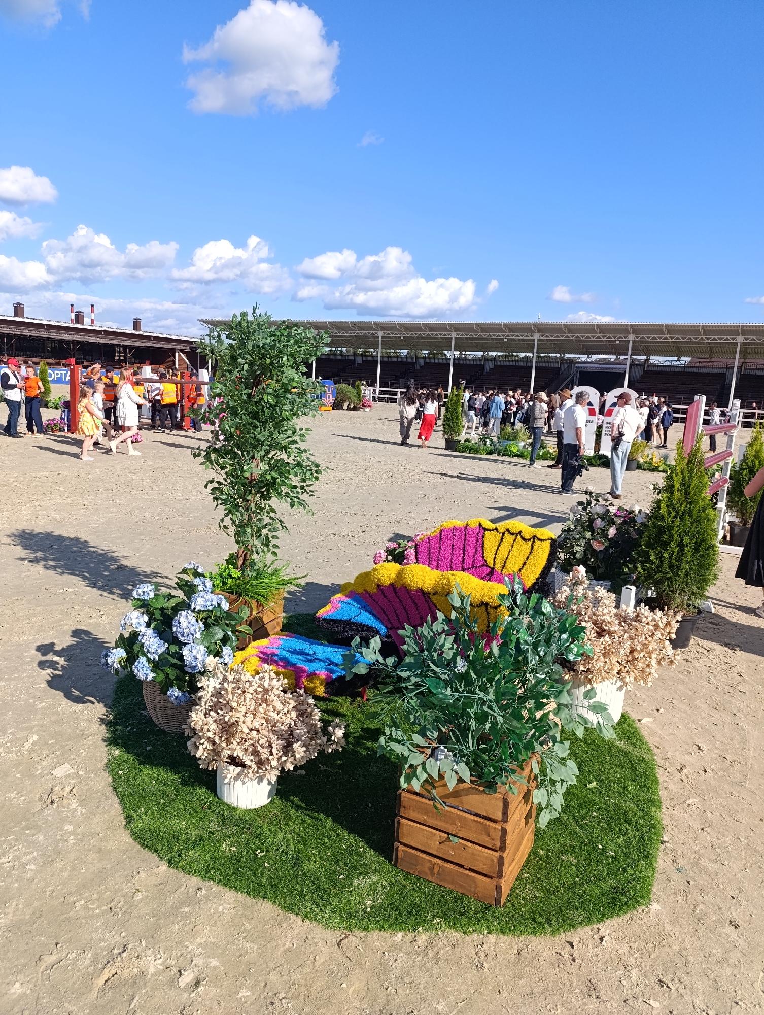 This media file captures an outdoor event with numerous people gathered. The scene appears to be a sandy arena, possibly at a equestrian facility, with covered grandstands visible in the background. The weather is sunny with scattered clouds, suggesting a pleasant day.
In the foreground, a decorative arrangement of flowers and greenery is prominently displayed. This includes a large, colorful butterfly made of flowers, with vibrant yellow, pink, and blue petals. Around the butterfly are potted plants, including blue hydrangeas in a woven basket, dried light-colored flowers in a white pot, and lush green foliage in a wooden planter. A faux tree also stands nearby.
The subjects in the image are a diverse crowd of individuals, dressed in casual summer attire. Some are walking, while others appear to be observing or participating in an event. A few people are positioned near the foreground floral display, while a larger group congregates in the middle ground, closer to the grandstands. Children are also visible among the attendees.
The overall atmosphere seems festive and relaxed. The bright sunlight casts shadows, indicating it is likely midday. The presence of grandstands and an arena suggests a competition or exhibition is either taking place or about to commence. Visible text in the image includes "OPTА" on a banner in the background, which might indicate a sponsor or part of the event's name. The location is identified as Ivanovskoe, Russia.