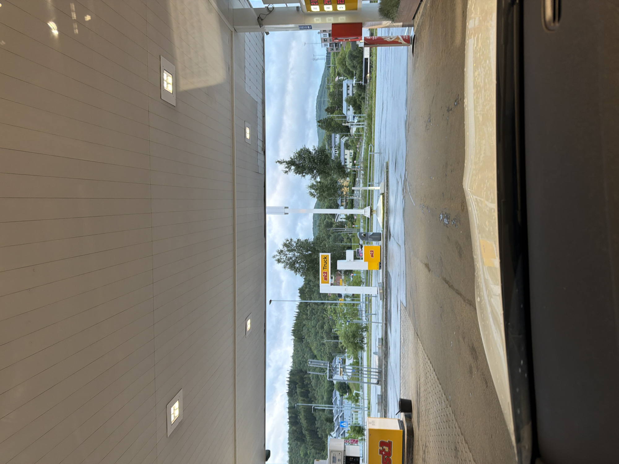 The image shows a view from inside a vehicle looking out from under a gas station canopy. The canopy is white with recessed lighting fixtures. To the left, a yellow box with the logo "st1" is visible. In the center of the frame, there is a white pole with a yellow sign that says "st1 Truck". Further back, a gas pump is visible. Beyond the gas station, the landscape includes trees and buildings, suggesting a semi-urban or suburban setting. In the background, a hill covered in dense green trees rises, and the sky is overcast. The ground in front of the gas station appears wet, indicating recent rain. This scene is located at the ST1 gas station in Harstad, Norway.