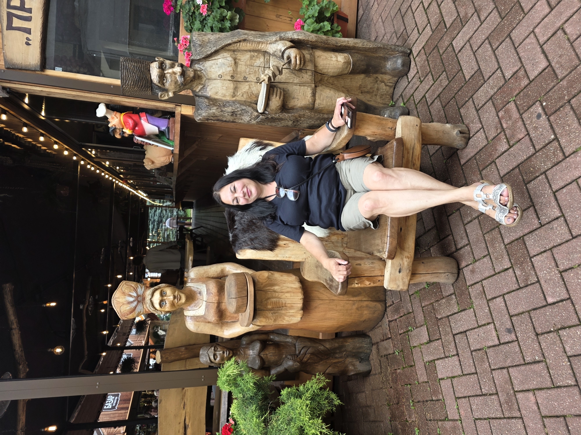 In the city of Estosadok, Russia, a woman with dark hair and glasses sits on a large wooden chair. She is wearing a black t-shirt and khaki shorts, with silver sandals on her feet. To her left are two wooden sculptures: a female figure holding a round object, and a smaller, nude female figure. To her right stands a large wooden sculpture of a man wearing a hat and fur-lined coat, holding an object in his hand. The scene is set on a brick-paved area, with greenery and wooden structures in the background, suggesting an outdoor restaurant or market setting. The overall lighting suggests it is daytime.