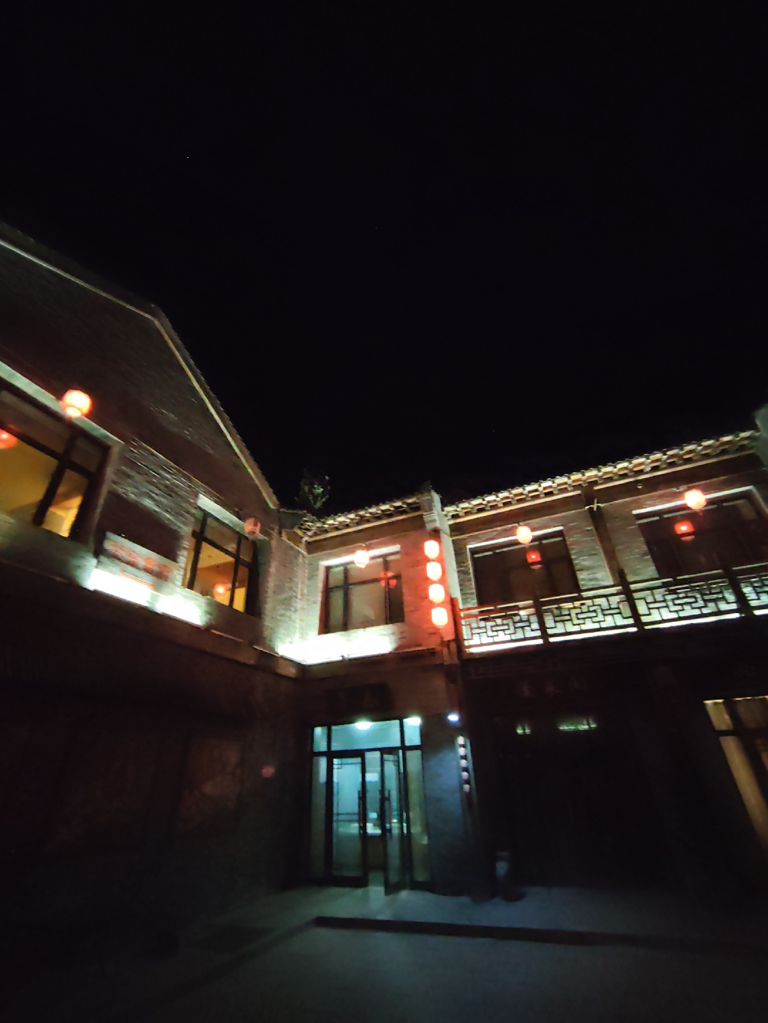 This is a nighttime exterior shot of a building with traditional Chinese architectural elements, located in Lindian, China. The building features brick walls and a tiled roof. Several red lanterns are illuminated and hung outside, casting a warm glow. The windows on the upper floor are lit from within, revealing yellow light. A decorative railing with a geometric pattern runs along the upper facade. In the lower part of the image, there is a doorway with glass doors, which appear to be open, leading into a brightly lit interior. The ground in front of the building is paved. The sky above is dark and clear, with a few faint stars visible.