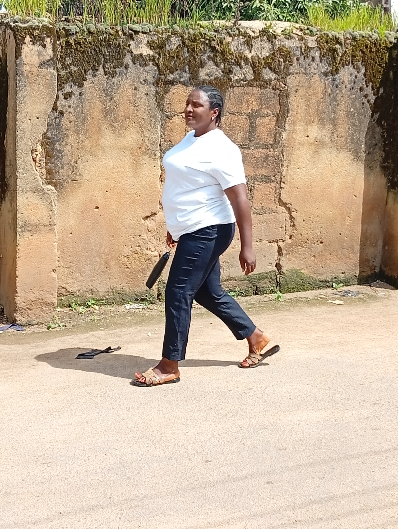 A full shot shows a woman walking on a paved surface in front of a weathered, textured, light brown wall covered in moss and a few green plants at the top. The woman is wearing a white t-shirt, dark blue pants, and brown sandals. She is holding a black umbrella in her left hand, and a black object, possibly a folded garment, lies on the ground behind her left foot. The scene is outdoors, and the lighting suggests it is daytime. The image appears to be taken in Jos, Nigeria, a city known for its natural beauty and historical structures.