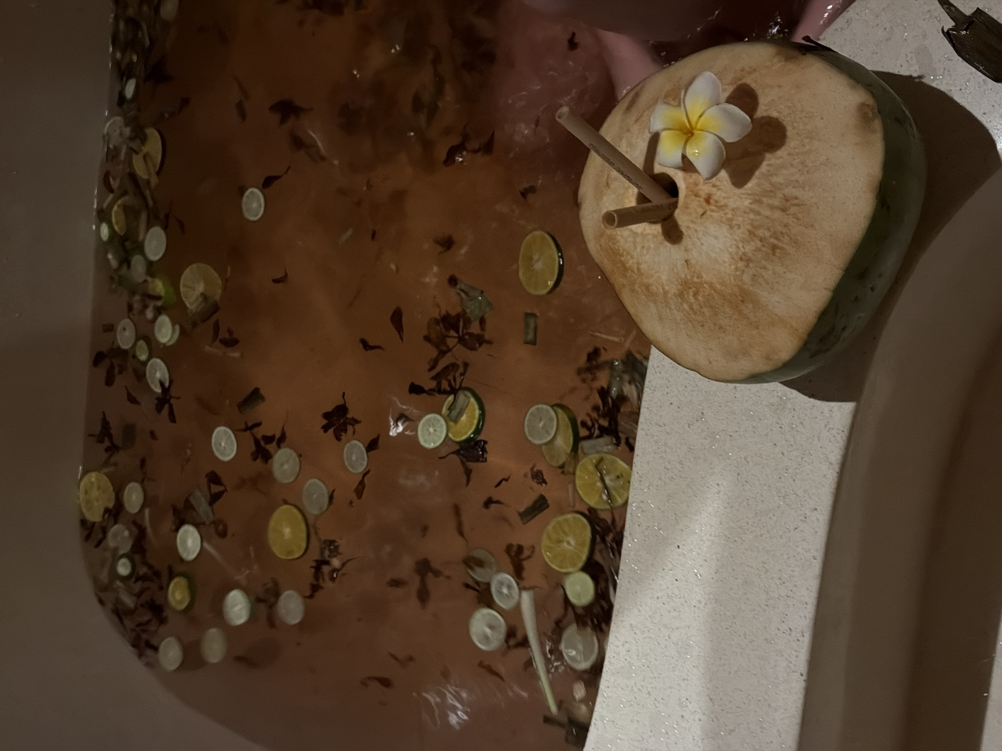 This is an overhead shot of a bathtub containing reddish-brown water, filled with various natural elements, alongside a tropical drink.
The bathtub water is murky and dark, with numerous thin, circular slices of citrus fruits, primarily limes and lemons, floating on the surface. Interspersed among the citrus slices are many dark, dried leaf fragments and some longer, slender pieces of plant matter, possibly lemongrass or cinnamon sticks, creating a natural, aromatic bath.
On the right edge of the tub, a young, green coconut, with its top neatly cut open, rests on a white, speckled surface. Two straws, one light-colored and one resembling bamboo, are inserted into the coconut. A delicate white and yellow plumeria (frangipani) flower is placed near the straws on the coconut, adding to the tropical aesthetic.
A person's arm or leg is partially visible, submerged in the upper right quadrant of the bathwater, suggesting someone is currently enjoying the bath. The lighting is dim and warm, casting soft shadows, indicative of an indoor setting, likely in the evening or a low-light environment, which enhances the relaxing ambiance.
The fresh coconut and plumeria flower are strong indicators of a tropical location, consistent with North Kuta, Indonesia. The overall scene suggests a luxurious, relaxing, and spa-like experience. There is no visible text in the image.