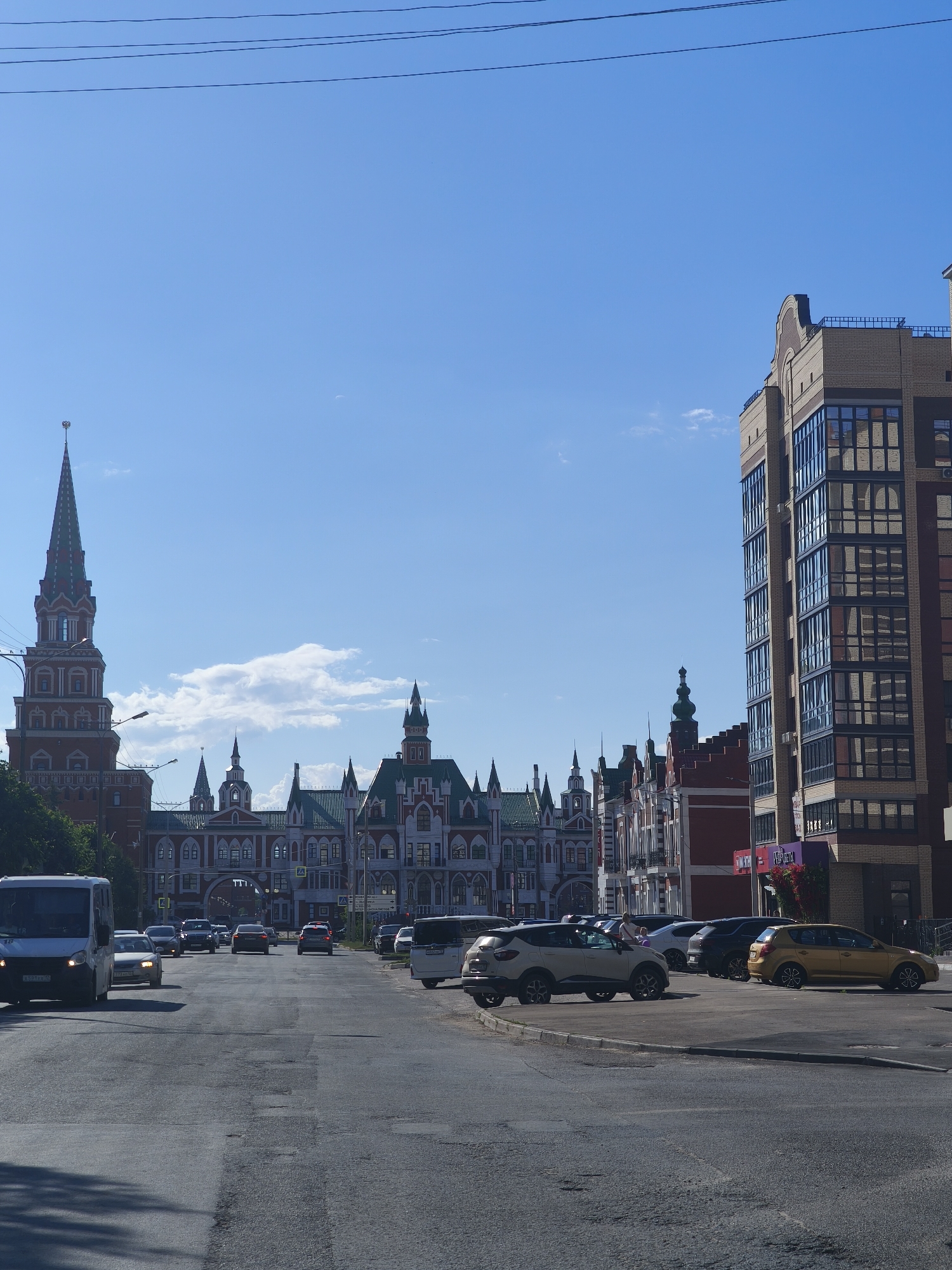 The image depicts a street scene in Yoshkar-Ola, Russia, with several buildings visible. On the left, a tall, red, spire-topped tower stands out, characteristic of the National Art Gallery. To its right, a long, ornate building with a green, multi-gabled roof and red and white accents stretches across the middle ground, likely the National Art Gallery itself or a connected structure. Further to the right, a modern, multi-story brick building with large, dark-framed windows dominates the right side of the frame. The street in the foreground is asphalt and appears to be moderately trafficked, with several cars and a white van visible. The sky is clear and blue, with a few wispy clouds. The scene suggests a sunny day in a city that blends historical and contemporary architecture.