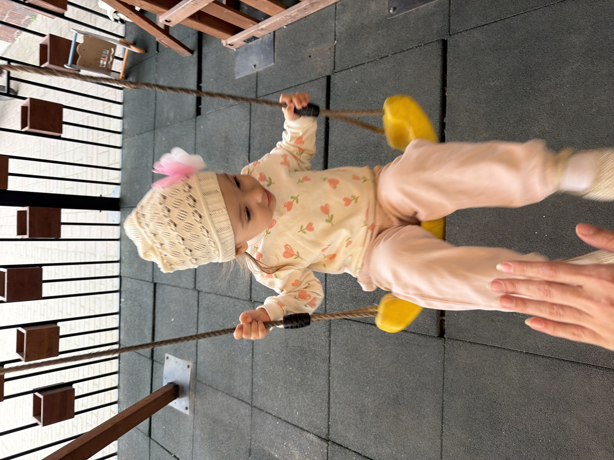 A young child, likely a toddler, is swinging on a yellow swing set on a grey rubberized playground surface. The child is wearing a white knitted hat with a pink flower, a long-sleeved shirt with a floral pattern, and pink pants. The child is holding onto the swing rope with their right hand. In the foreground, a person's left hand is visible, extended towards the swing. The background shows a black metal fence with wooden planters attached to it, and part of a wooden play structure. The environment appears to be an outdoor playground. There is no information to determine the specific location within Ufa, Russia.