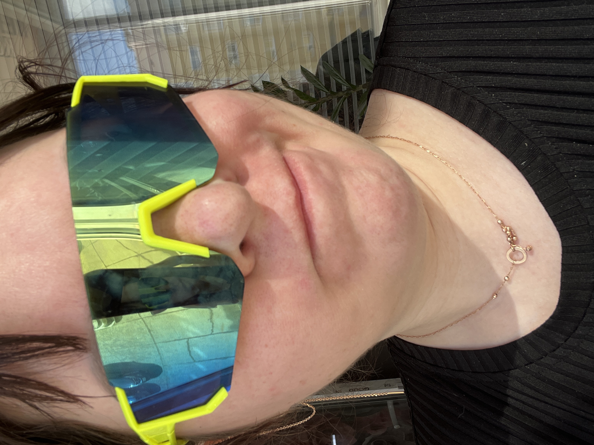 The image presents a close-up portrait of an individual, identified as being in the city of Noviy, Russia. The subject, an adult appearing female, is framed from the upper chest to just above the forehead. She has dark brown hair and is wearing large, sporty sunglasses with distinct neon yellow frames and highly reflective, blue-green iridescent lenses that obscure her eyes. The reflections in the lenses show an interior setting, with the left lens reflecting what appears to be a patterned surface, and the right lens reflecting a window with horizontal blinds and an exterior light-colored building or wall. The subject exhibits a slight upturn at the corners of the mouth, indicating a minimal smile. Around her neck, a delicate rose gold-colored chain necklace with small spherical beads and a central circular charm, from which a smaller ring and a bead are suspended, is visible. A gold-toned chain earring is partially visible on the subject's left side. She is dressed in a black top with vertical ribbing. The background to the subject's right features a window with horizontal blinds, providing a partial view of an exterior light-colored structure and a green leafy plant.