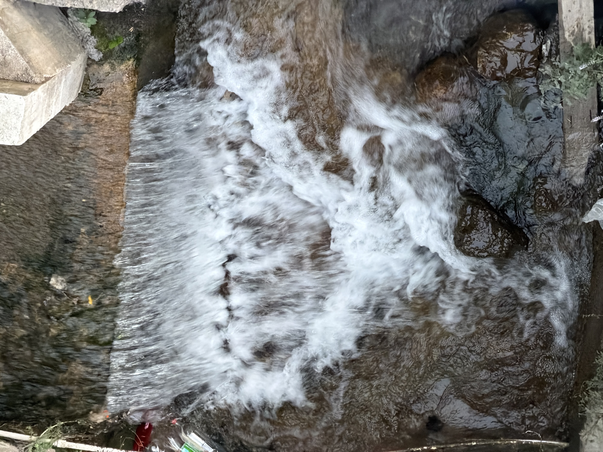 The image is an overhead, close-up view of a small, man-made waterfall flowing into a shallow channel. The water is a bright white and foamy as it cascades over a rough, dark brown ledge, suggesting it's a rocky or concrete structure. Below the waterfall, the water is more diffused and darker, with visible rocks submerged in the channel. The channel itself appears to be constructed of dark, wet earth and stone. The surrounding environment is not clearly visible, but hints of green vegetation can be seen at the edges, particularly to the right and bottom of the frame. There are also some pieces of debris, including what appears to be plastic, in the water and on the banks. The scene is devoid of people. The lighting is natural and appears to be from above.