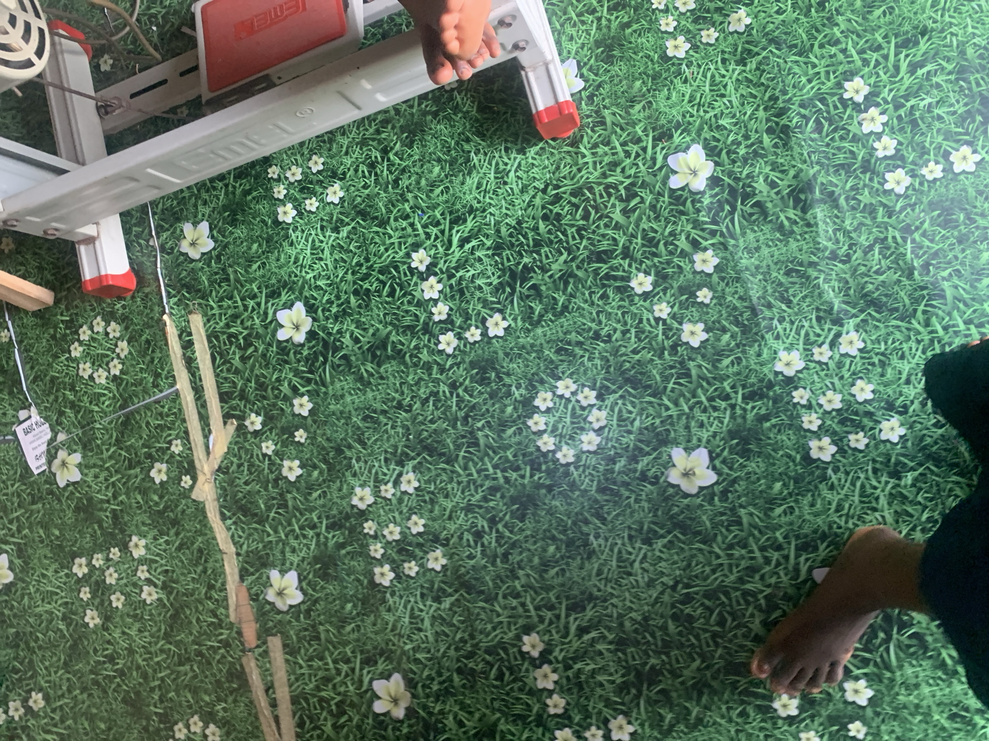 The image shows a close-up, overhead view of a green carpet with a printed grass and flower design. A person's dark-skinned feet are visible in the lower left and right of the frame. In the background, a sewing machine, primarily white with red accents, is partially visible on the right side. A label that reads "BASIC HUG" and "PRINTED" is partially visible in the upper center of the image. The scene appears to be indoors, possibly in a home or workshop. The specific location of Jauro Yinu, Nigeria, cannot be determined from the visual information in the image.