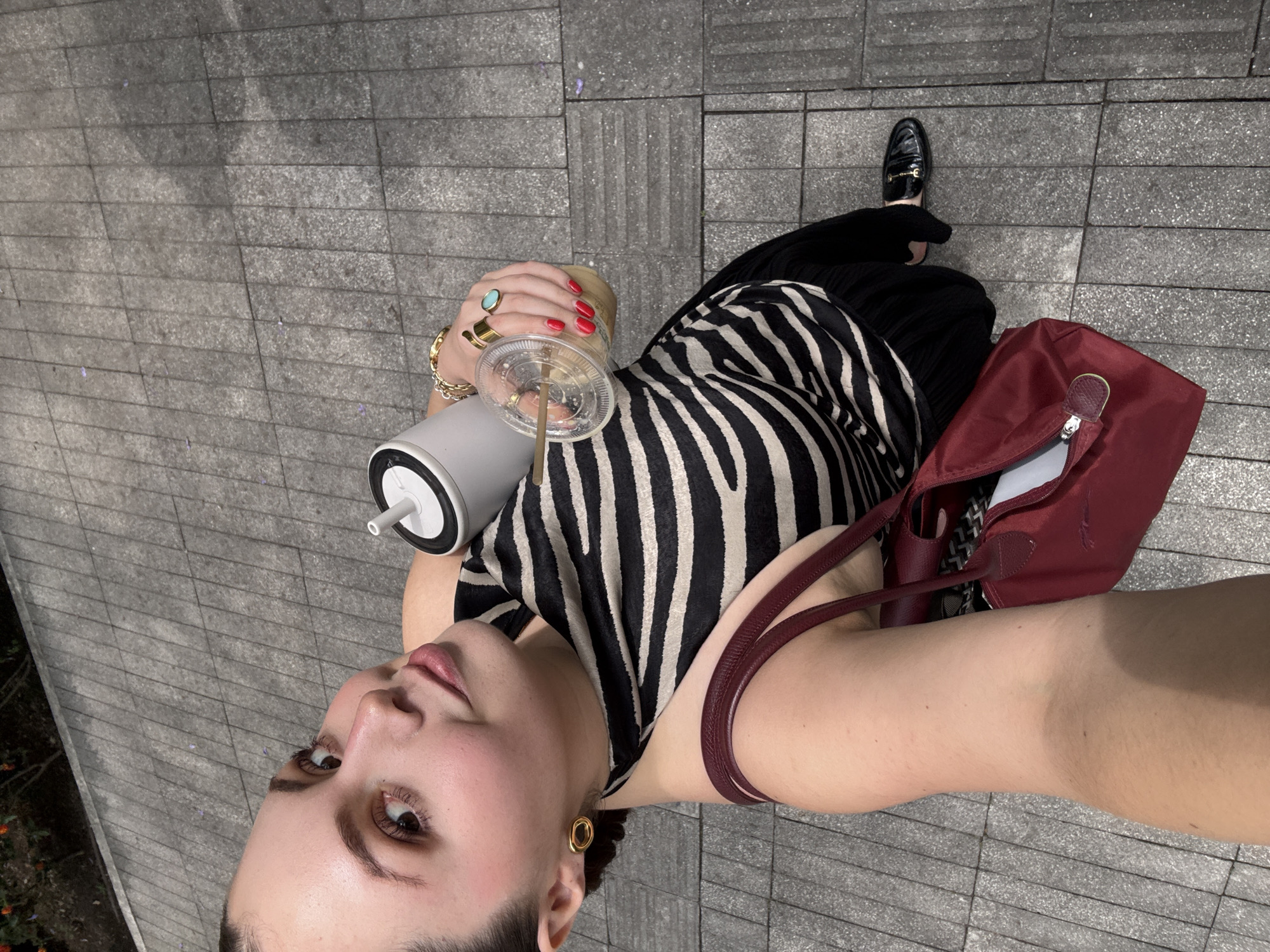 A woman stands on a paved surface, likely a sidewalk, holding a clear plastic cup with a straw and a white thermos. She is wearing a zebra-striped top, black pants, and black loafers. Her right hand is adorned with multiple gold bracelets and rings, and her fingernails are painted red. She also has gold hoop earrings. A burgundy-colored tote bag hangs from her shoulder. The background consists of grey paving stones.
The image was likely taken in Mexico City, Mexico, indicated by the scattered jacaranda flowers on the ground. Jacaranda trees are common in Mexico City, particularly in spring, and their purple flowers are often seen on sidewalks and streets.