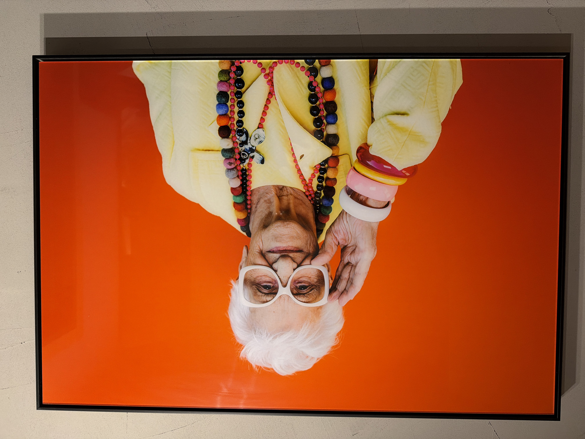 A photograph of an elderly woman with white hair and large white-rimmed glasses, wearing a yellow jacket and colorful beaded necklaces and bracelets, is framed in a black frame and hung on a wall. The background is a solid orange color. The photo is upside down. The wall is light grey. It is difficult to determine if the photograph is in Sofia, Bulgaria.