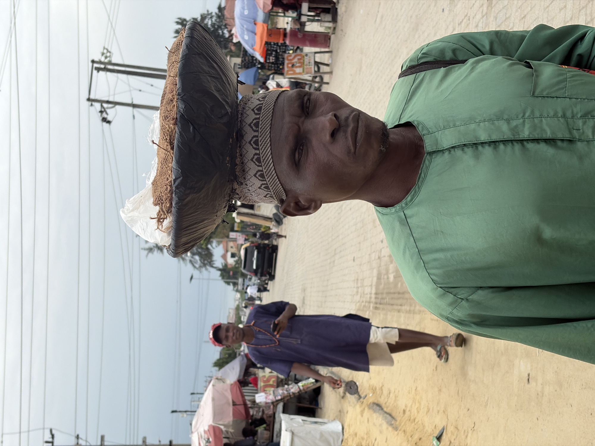 In Igboefon, Nigeria, a man in a green shirt and patterned cap balances a large tray covered in a black plastic bag on his head. The tray contains brown, fibrous material. In the background, another man in a blue shirt and light-colored shorts stands on a sandy surface, near stalls with red and white canopies. A street with cars and pedestrians is visible in the distance, beneath an overcast sky. Utility poles and lines are also present.