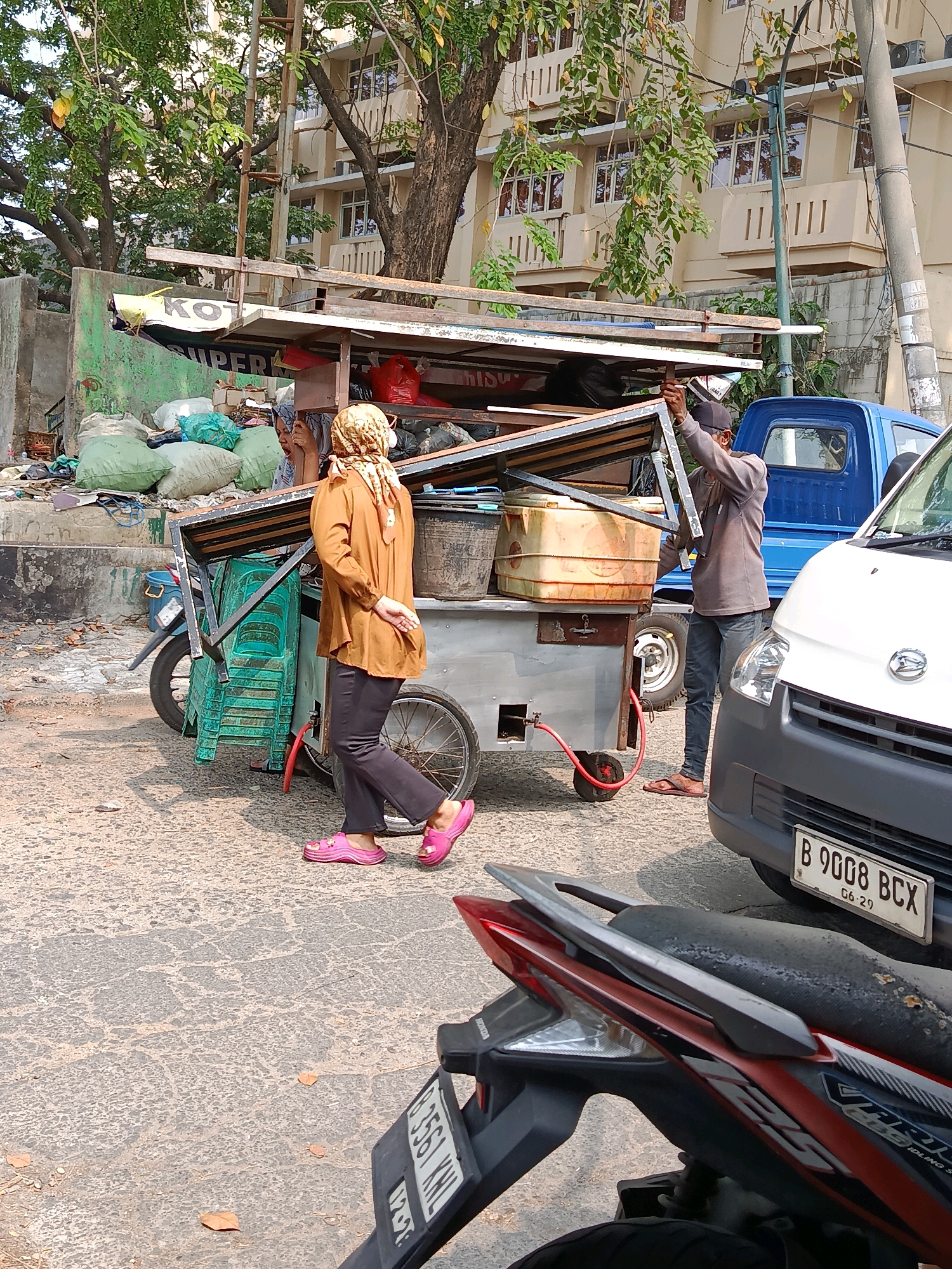 In Bekasi, Indonesia, a woman wearing a brown long-sleeved shirt, dark pants, a headscarf, and pink slip-on shoes walks past a food cart. The food cart, which appears to be on a street, is laden with various items including stacked green plastic chairs, a large translucent yellow container, and a red bag. A man wearing a grey long-sleeved shirt, dark pants, a black baseball cap, and flip-flops is standing by the cart, holding onto a metal frame. A blue pickup truck is parked behind the cart, and a white vehicle is in the foreground. The ground is paved with asphalt, and debris is visible on the left side of the image, alongside a concrete wall and vegetation. A motorcycle is partially visible in the bottom right of the frame.