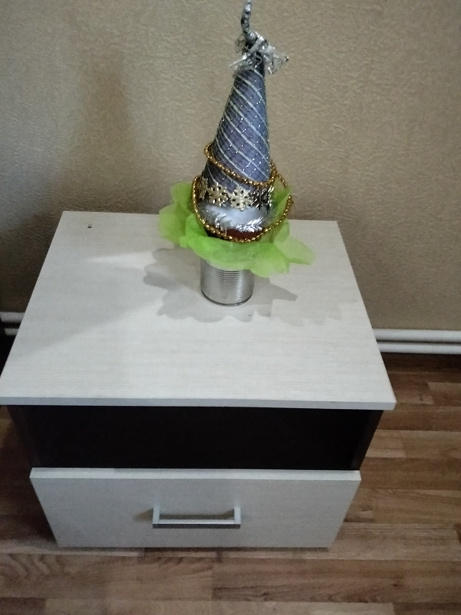A conical decorative object, resembling a Christmas tree, stands on a light-colored wooden tabletop. The object is adorned with silver and gold beads, and a light green fabric is draped around its base. It is supported by a silver-colored metal can. The tabletop is part of a multi-level furniture piece with a dark brown section and a light-colored drawer with a silver handle. The background is a textured, beige wall. The floor is made of light brown wooden planks. This scene appears to be indoors, likely in a residential setting in Nizhnedonskoy, Russia. There are no people or animals visible in the image.