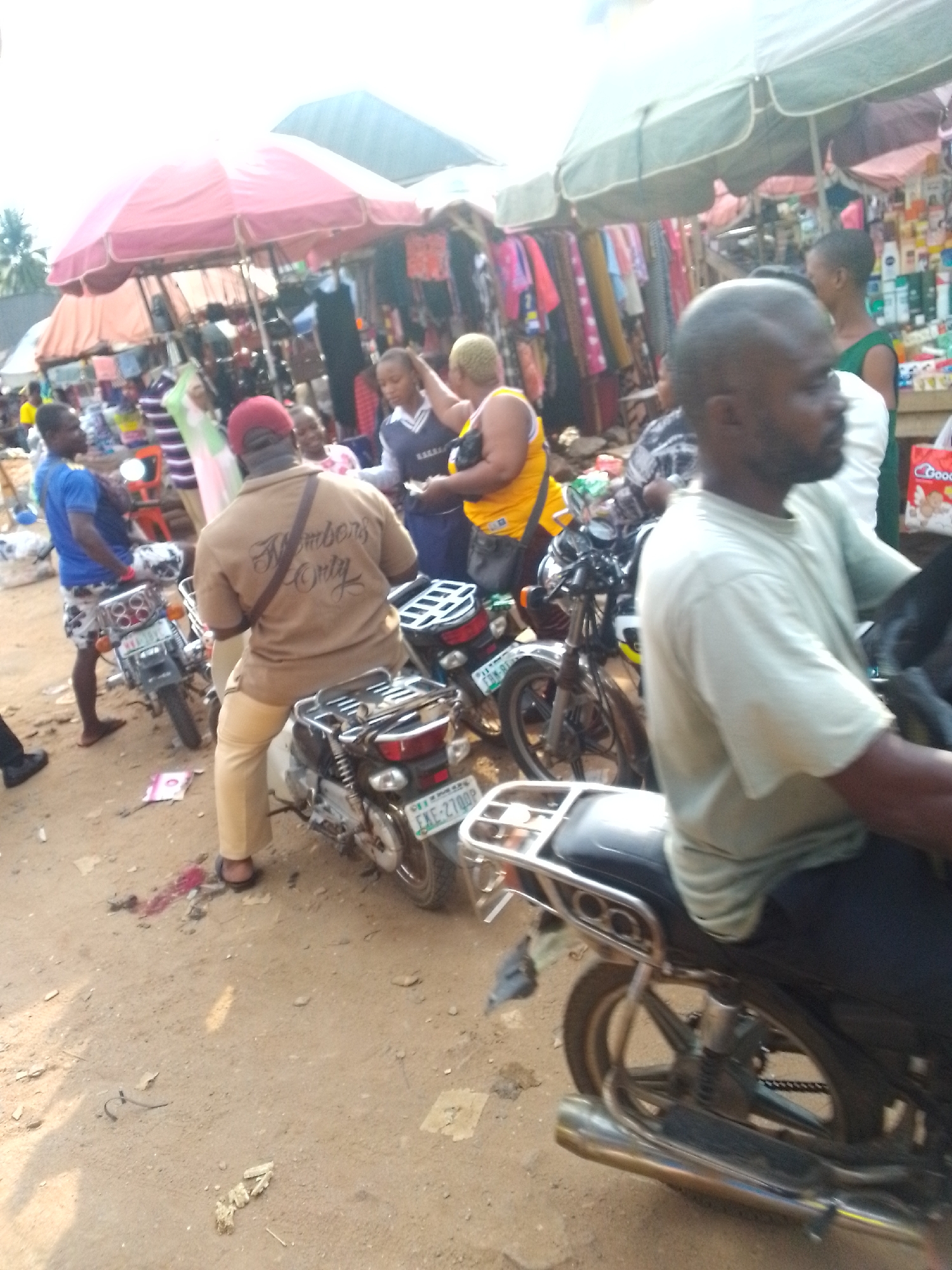 The image shows a bustling marketplace in Orlu, Nigeria. Several people are visible, including men on motorcycles. Some are shopping, others are interacting with each other. Clothing and other goods are displayed under makeshift shelters with umbrellas. The ground is dirt. License plates on the motorcycles are visible.