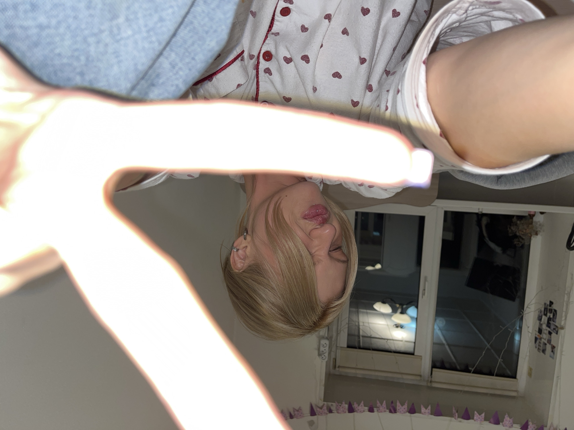 The image shows a person with blonde hair, wearing pajamas with small red hearts on a white background. Their right eye is squinted shut, and they are sticking their tongue out slightly. The person has a hand with fingers extended towards the camera, obscuring the view. In the background, there is a window with a view that includes another building and lights. There is also a string of purple crowns along the wall. Small photographs are taped to the wall to the right of the window.
Given the location provided, it can be deduced that the photo was taken somewhere in Chaikovsky, Russia.