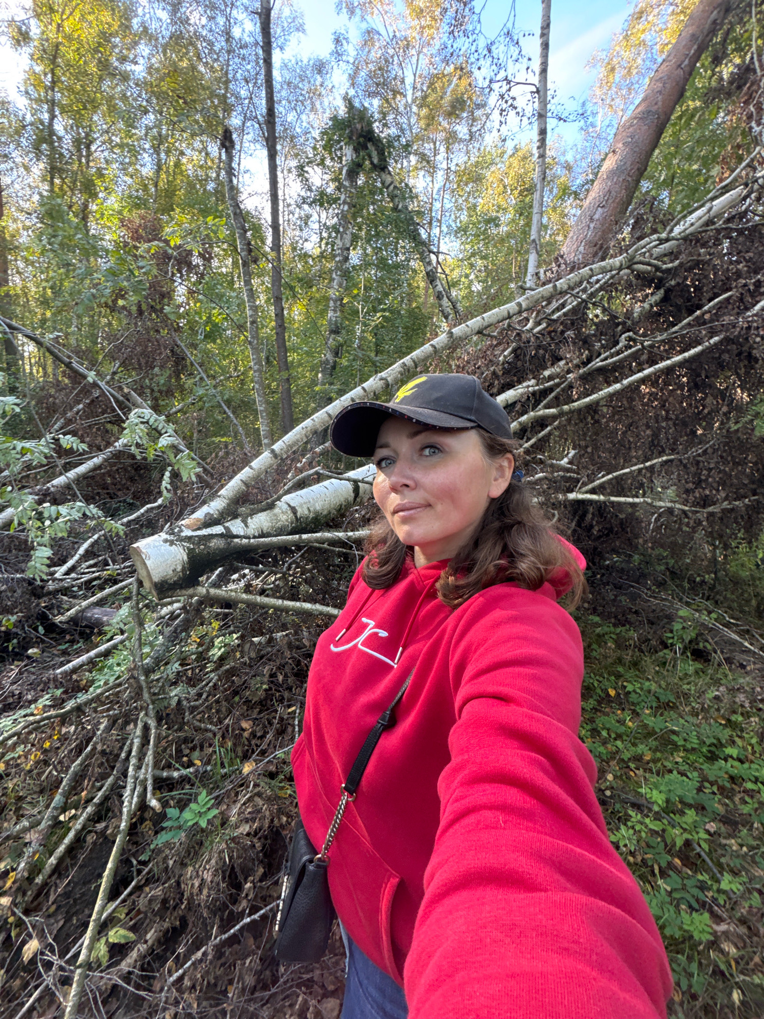 A woman in a red hoodie and a black baseball cap is taking a selfie outdoors. She is standing in a forest with a fallen tree and branches behind her. The leaves on the trees are green and yellow, suggesting it is autumn. The sun is shining through the trees, casting dappled light on the scene. The woman is looking at the camera with a slight smile. She is wearing a small black purse with a chain strap.