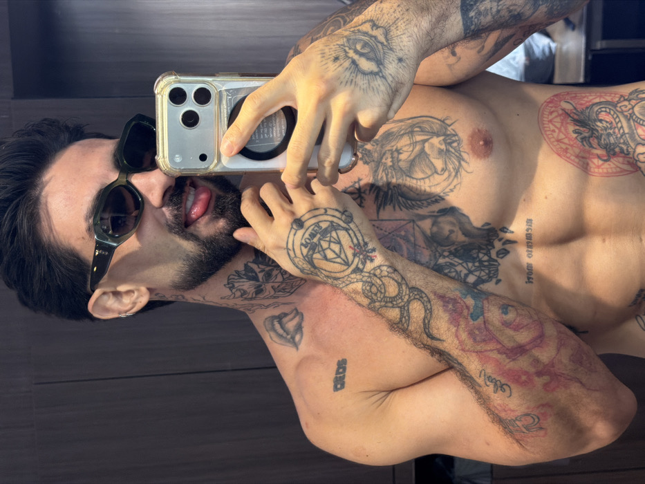 A heavily tattooed male individual, shirtless, is taking a mirror selfie indoors in Bangkok, Thailand. He has dark hair, a full beard, and wears dark oval sunglasses with a silver hoop earring in his left ear. His left neck displays tattoos of lips with a tongue and a split face (half demonic, half human). The left shoulder features the tattoo "DEOS". Extensive tattoos cover his arms, chest, and torso, including a snake, geometric patterns, a large skull, a dragon on the left pectoral, and a red-inked dragon within a geometric frame on his right lower rib cage. His left hand has a circular design with a diamond and the word "LOVED", while his right hand features an eye with radiating lines. He possesses a muscular physique with visible abdominal muscles. The subject holds a clear-cased iPhone with a MagSafe ring in his right hand, using it to capture the image. His left hand is raised, partially obscuring his chin. He is sticking his tongue out slightly. The background consists of dark wooden panels or furniture.
