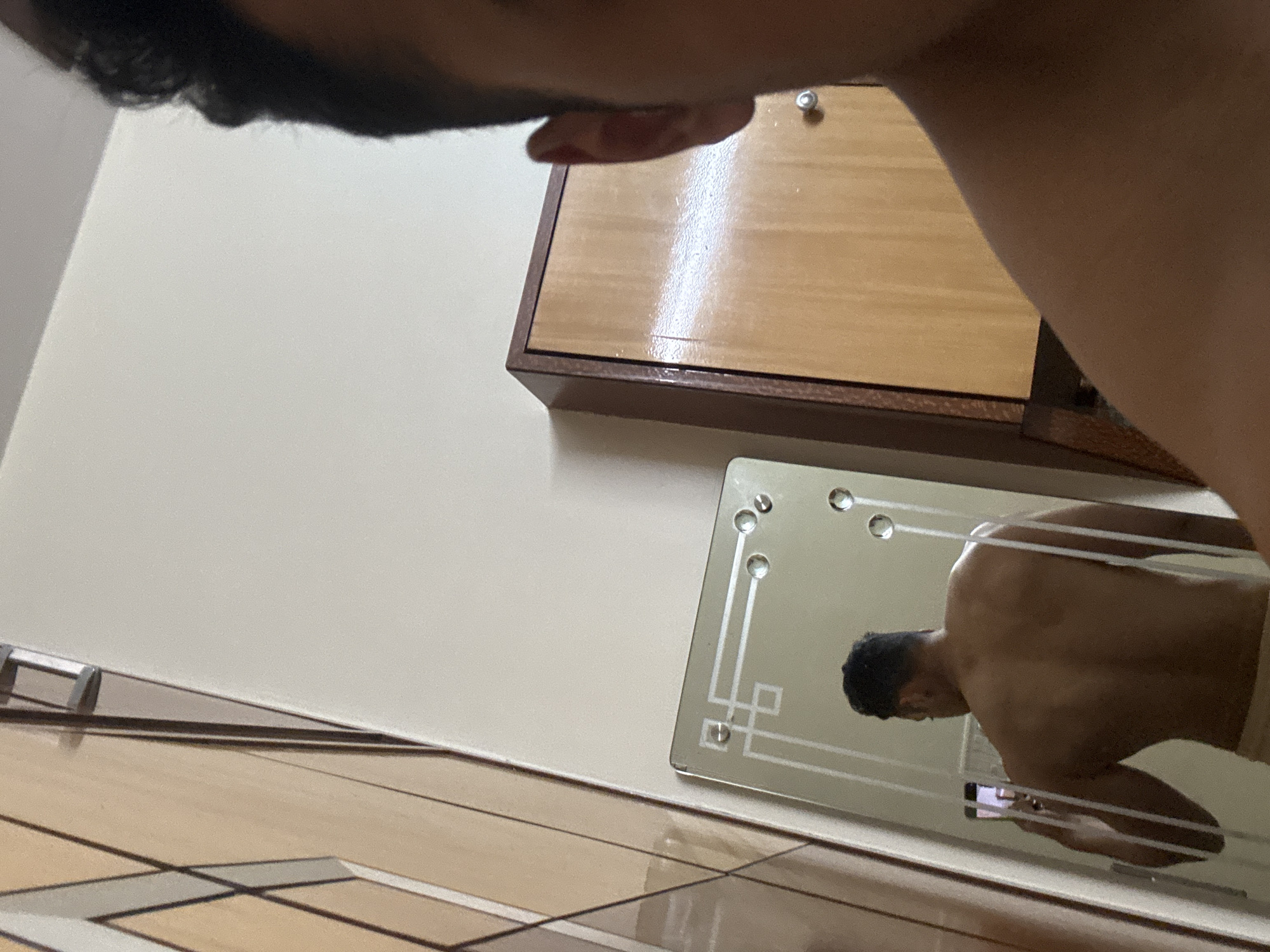 The image depicts a man taking a selfie in a mirror. The man is only partially visible in the frame, with his head and shoulder appearing in the upper left corner of the image. A wooden cabinet hangs on the wall above the mirror. The reflection in the mirror shows a shirtless man, presumably the same one taking the photo, holding a phone. The floor appears to be tiled. The location is unknown and cannot be inferred to be Kolkata, India, from the image alone.