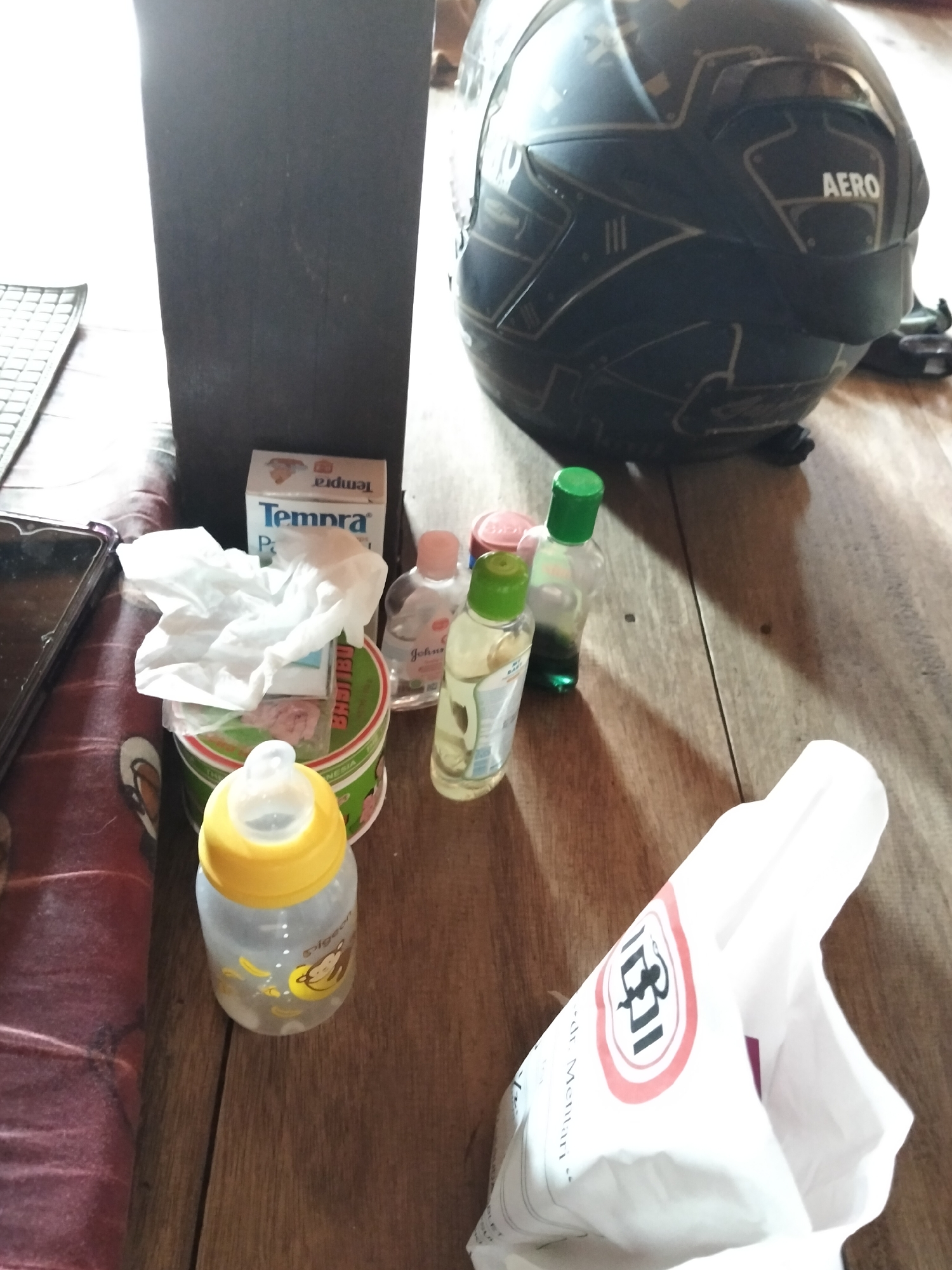 This is a close-up, high-angle, indoor photograph of various items on a wooden surface. The items are arranged in a cluttered manner. Prominently featured is a black motorcycle helmet with gold accents and "AERO" branding. To the left of the helmet is a dark, rectangular object, possibly a speaker or cabinet.
In the foreground and midground, there are several baby-related items. These include a baby bottle with a yellow cap and cartoon monkey graphics, a small container of what appears to be baby powder or cream, a crumpled white tissue, and several small plastic bottles with green and pink caps, likely containing baby oil or lotion. One of these bottles has the "Johnson's" logo visible. A box labeled "Tempra P" is also present.
Further to the right, a white plastic bag is partially visible, with a red and black logo featuring a monkey-like figure and the text "IJN". The overall scene suggests a home environment, possibly a bedroom or living area. The lighting is somewhat diffused, creating shadows on the wooden surface.
The location in Setia Jaya, Indonesia, cannot be determined from the visual information provided in the image.