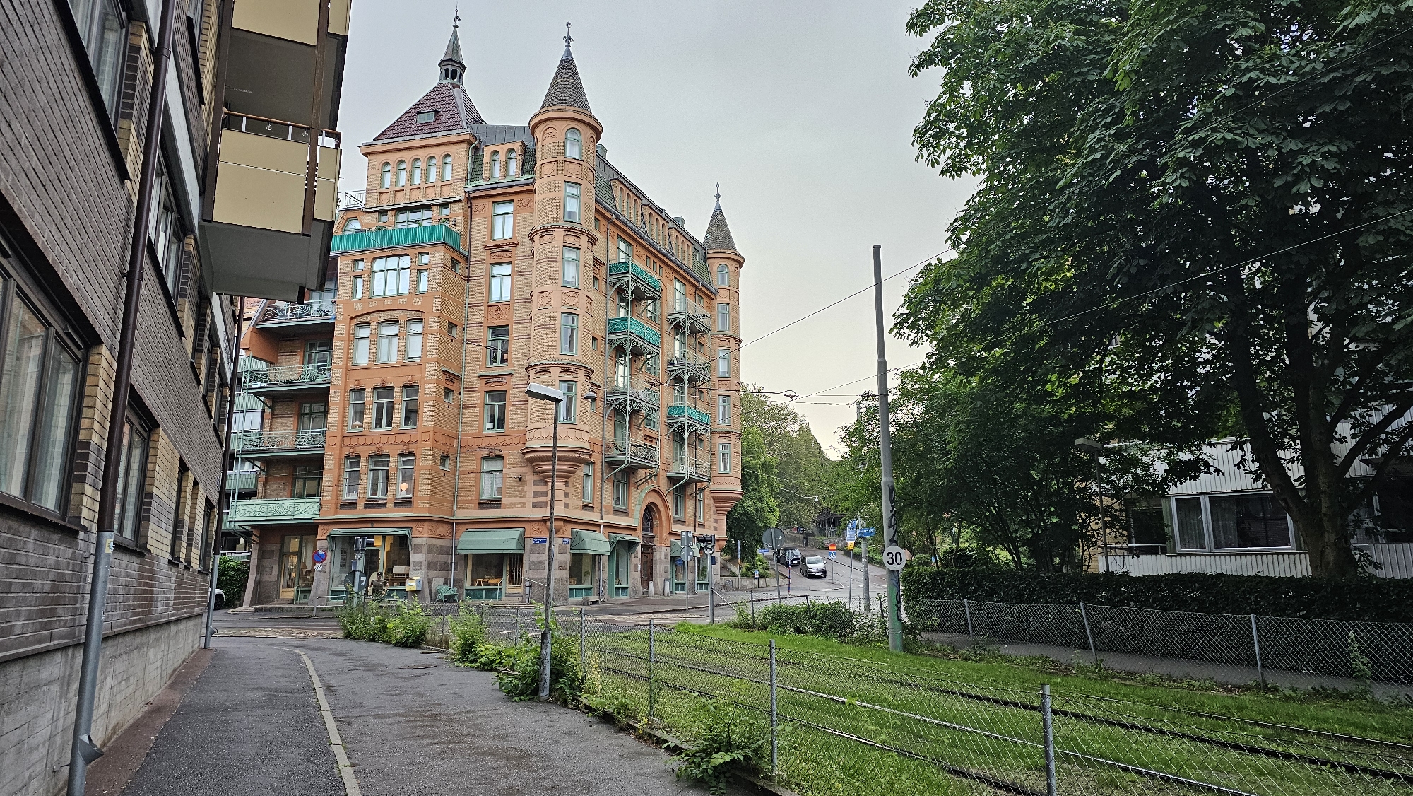 The image depicts a street scene in Gothenburg, Sweden, likely in an older, established neighborhood given the architecture. On the left is a modern brick building with multiple floors and windows. Adjacent to it is a tall, ornate, salmon-colored building with a red-tiled roof, turrets, and decorative green balconies. This building appears to be an apartment complex with ground-floor retail spaces. A street with tram tracks runs in front of the ornate building and extends into the background. Lush green trees line the right side of the street, partially obscuring a white, modern building with windows. The sky is overcast. The overall atmosphere is quiet and residential, with a few cars visible in the distance. A "30" speed limit sign is visible on a pole. The scene is a typical urban streetscape with a mix of older and newer architectural styles.
