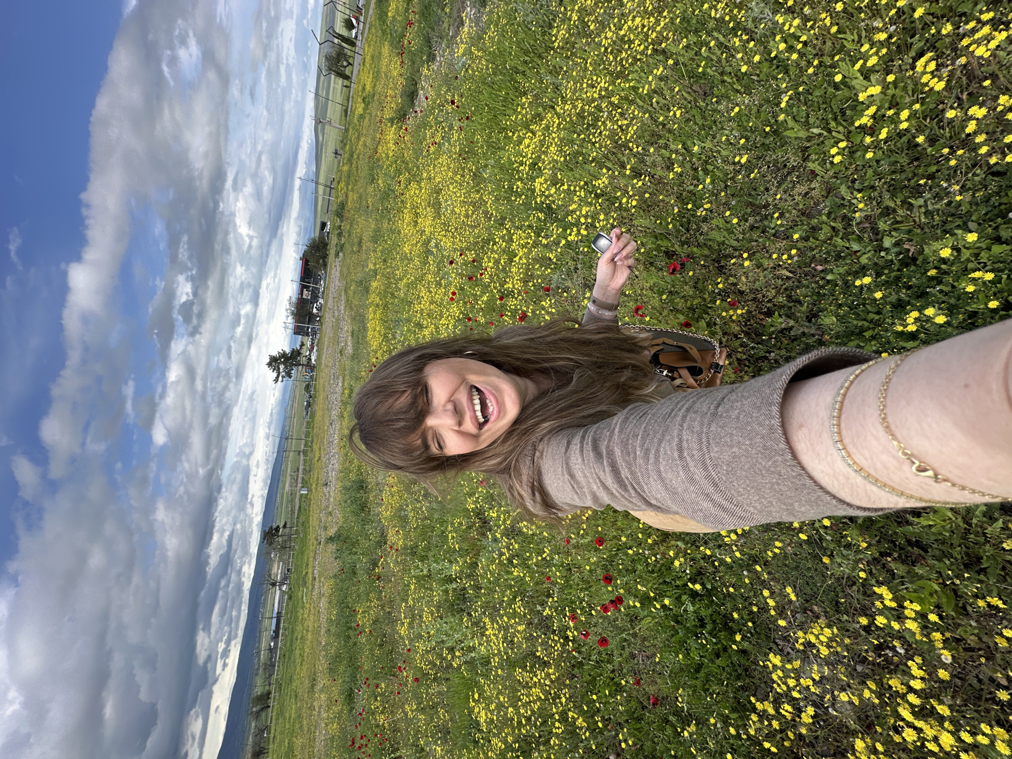 A woman is taking a selfie in a field of yellow and red flowers, under a partly cloudy sky. The location appears to be in Marneuli, Georgia, evidenced by the landscape and fence line in the background. The woman has brown hair and is wearing a tan sweater. She is smiling broadly. In her hand, she is holding a metallic object, possibly a camera. She is also wearing a gold bracelet on her wrist. The field stretches towards the horizon, and beyond the fence line, there are mountains in the distance.