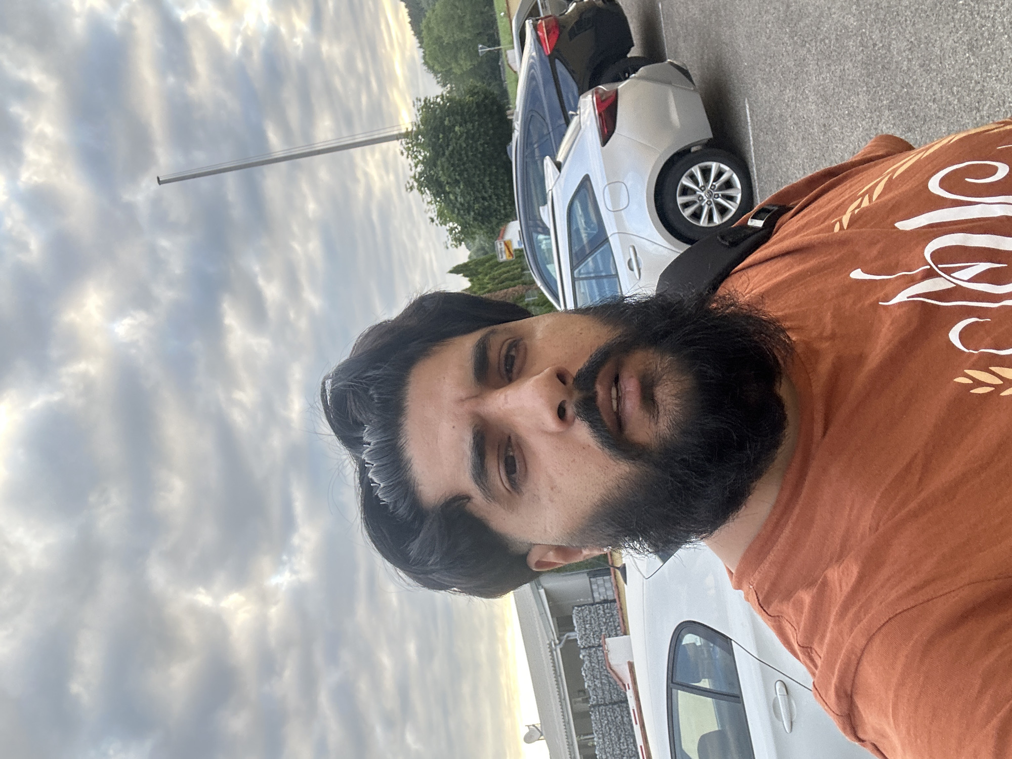 A close-up, slightly low-angle, outdoor selfie shows a man with dark hair and a beard. He is wearing an orange t-shirt. In the background, there are several cars parked on asphalt, and behind them, buildings and trees are visible under a cloudy sky. The image was taken in Mainburg, Germany.