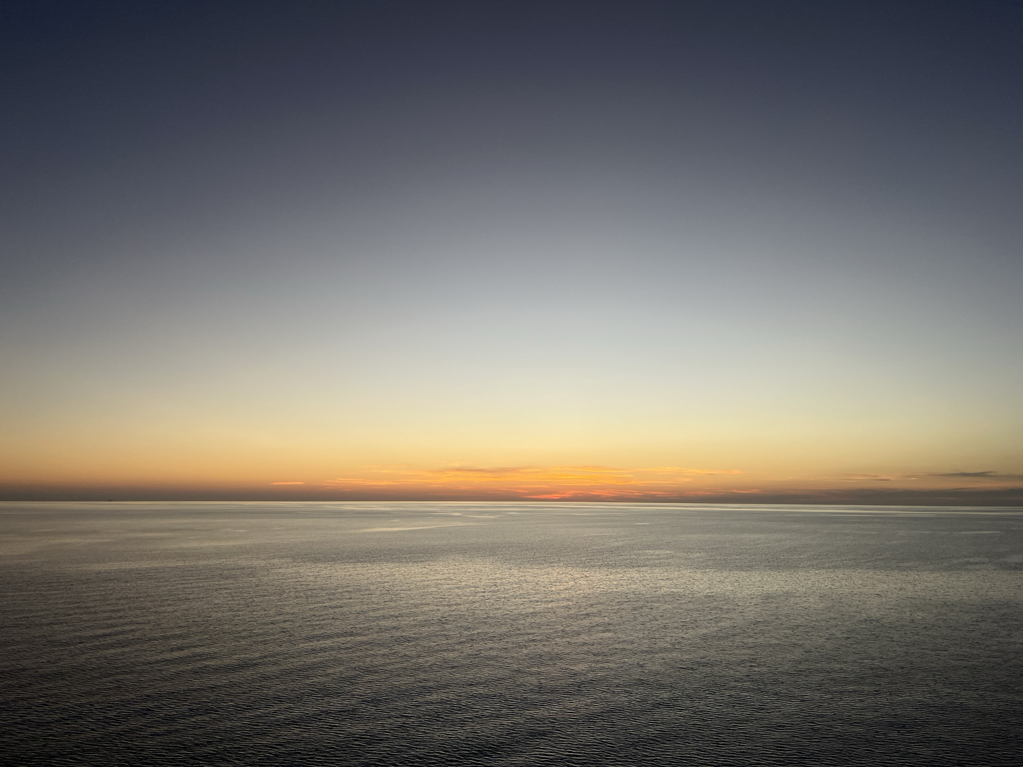 The image presents a serene seascape at sunset. The sky transitions from a deep blue at the top to a pale gradient of yellow and orange near the horizon. The sun's glow is visible through a thin layer of clouds on the horizon line, creating a warm color band. The sea reflects the colors of the sky, with subtle ripples across the surface creating a textured appearance. The location is at sea, with no discernible city or land features.