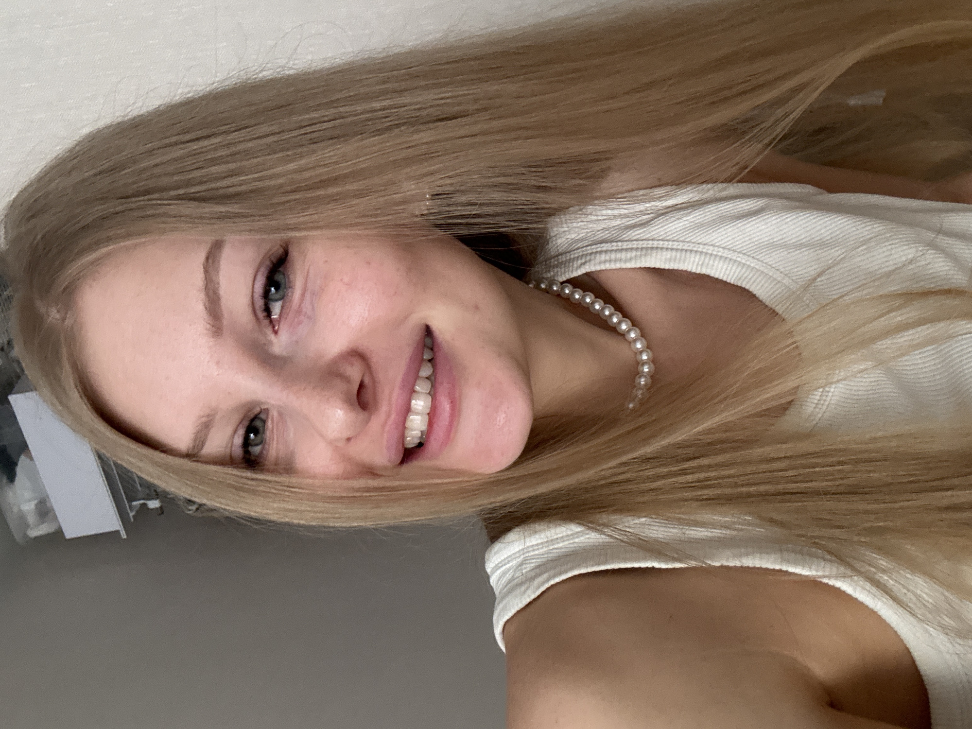 A young woman with long blonde hair is smiling at the camera. She is wearing a white top and a pearl necklace. The background is a neutral gray and white. There is no indication of location or time.