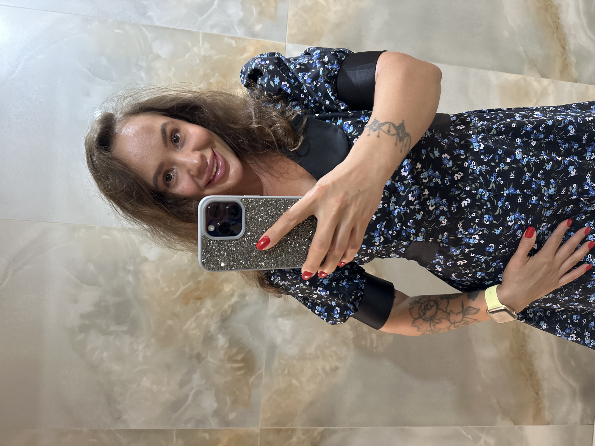 In Duvankul', Russia, a woman with medium-length brown hair is taking a mirror selfie. She is wearing a black dress with a floral print of blue flowers and puff sleeves. Her fingernails are painted red, and she has a watch on her left wrist. She is holding a smartphone with a sparkly case in her right hand. The background appears to be a wall with a marble-like texture.