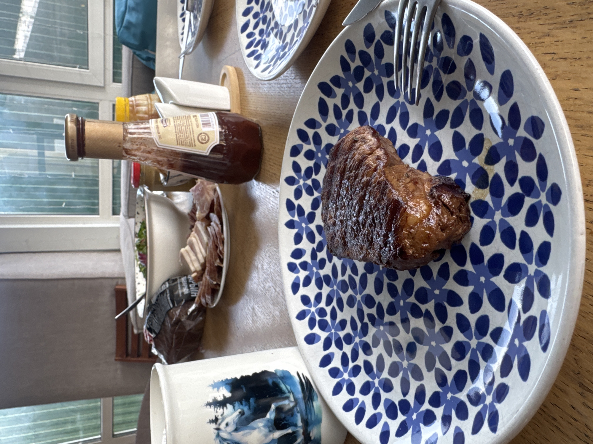 This image captures a tabletop setting prepared for a meal, likely in Ptichnoye, Russia, during daylight hours. No people are visible in the frame.
The central subject is a single portion of browned, cooked meat, possibly steak or roasted pork, resting on a round white ceramic plate. The plate is adorned with a repeating pattern of dark blue floral or stylized petal designs. A silver fork is placed at the top edge of this plate. Another identical plate is partially visible further up the wooden table.
To the left of the main plate, several condiments and side dishes are arranged. A tall, dark brown glass bottle with a golden cap and a beige label is prominent. Text on the label is mostly unreadable, but "BARBEQUE" might be partially discernible, suggesting it's a sauce. Behind it, a yellow squeeze bottle, likely containing mustard or another sauce, is partially seen. Further back, an oval white dish holds several slices of what appears to be bacon or cured deli meat. Adjacent to this, a dark plastic bag contains an unidentifiable food item, possibly bread, and a small white bowl with green salad is also present.
In the lower left foreground, a white mug features a distinct blue and white illustration of white horses in a wintery or forested landscape. The background of the scene reveals a large window with horizontal blinds, through which natural light enters, suggesting it is daytime. The overall impression is that of a casual home meal.