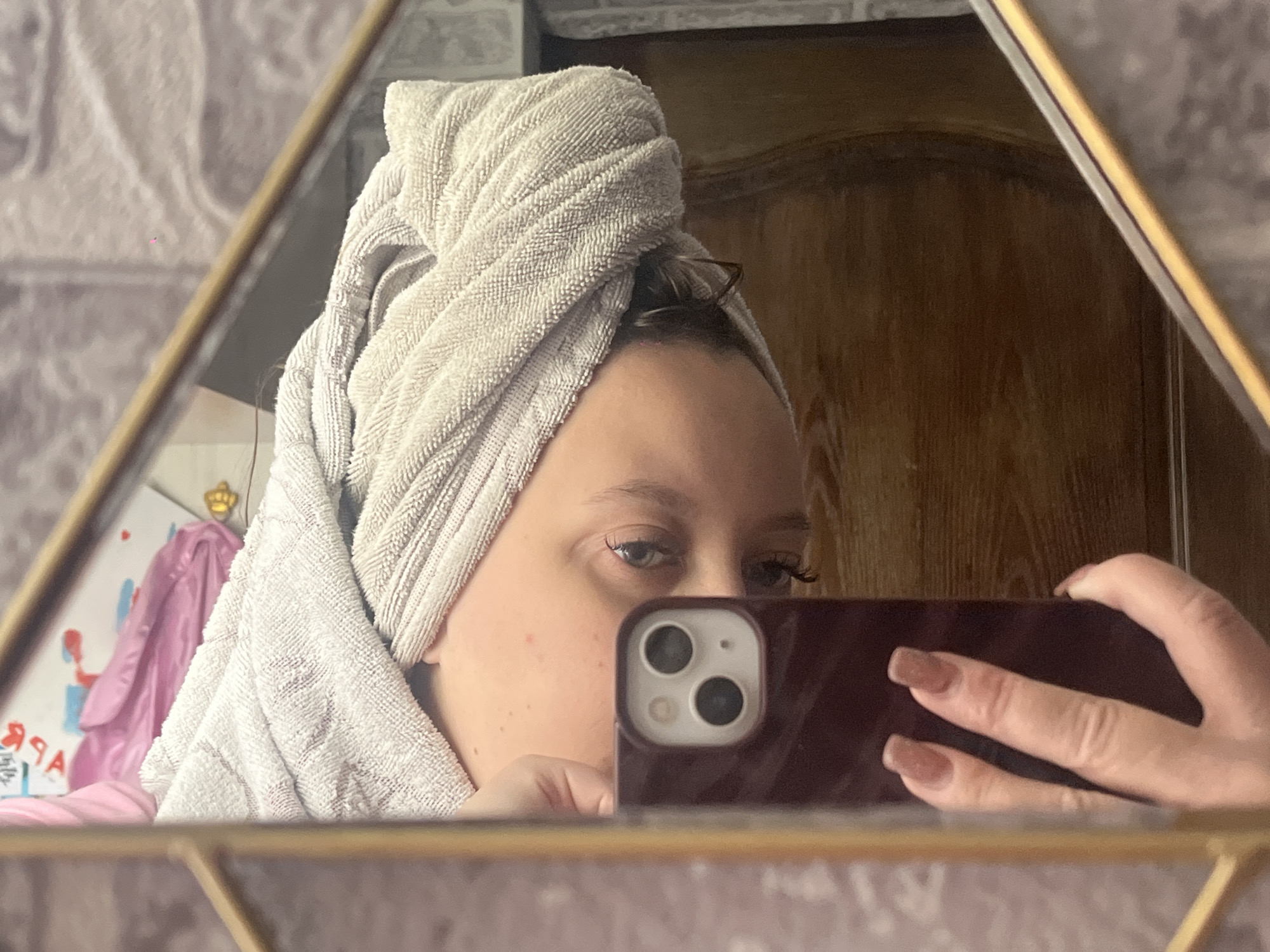 A person with a towel wrapped around their head is taking a selfie in a mirror. The person is holding a maroon-colored smartphone, partially obscuring their face. The background includes patterned wallpaper, a wooden door, and a drawing of a crown. The city of Novopokrovskaya, Russia, cannot be verified from the image.