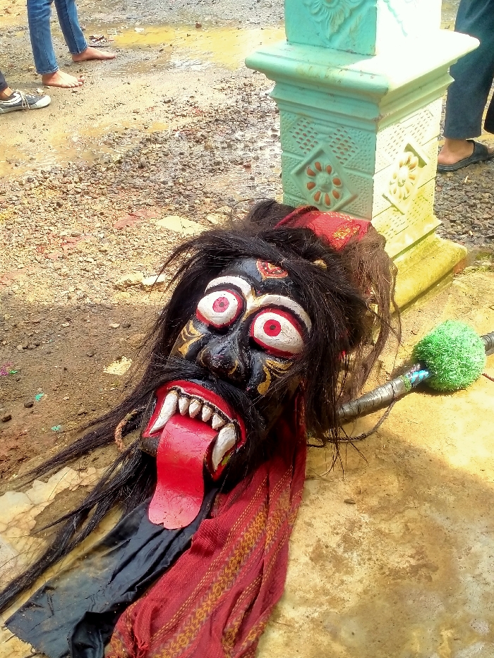 An elaborate traditional mask, depicting a stylized black face with wide white and red eyes, a red protruding tongue, and prominent white fangs, lies on a dirt and mud-covered ground in Gempolan, Indonesia. The mask features long, dark, matted hair-like strands extending from its head and is adorned with a red fabric draped around its lower portion, displaying a patterned golden design. Adjacent to the mask, to its right, rests a dark staff or club-like object with a spherical green, textured end. In the background, partially visible, are the lower legs and feet of at least two individuals; one pair is barefoot, standing on the wet, muddy ground, while another individual wears dark trousers and a dark slipper. A light-colored, possibly concrete, pillar with decorative carvings is also visible to the upper right of the mask. The ground varies between wet, reflective patches of mud and drier, rocky sections.