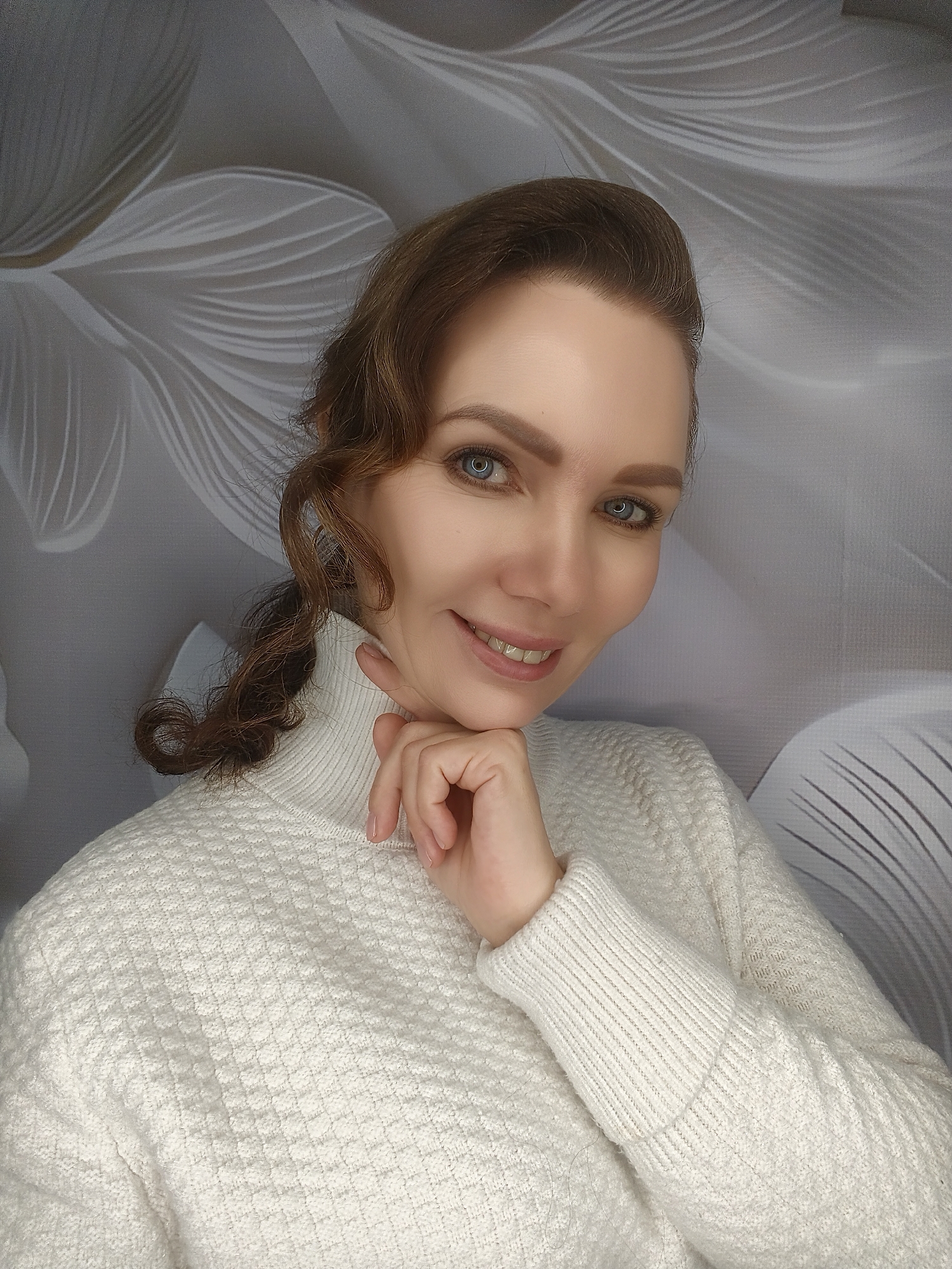 A woman with fair skin, brown hair styled with curls near her face, and blue eyes is smiling at the camera. She is wearing a textured white turtleneck sweater. Her hand is gently resting on her chin. The background appears to be a neutral-toned wall with a subtle floral pattern. The location is in the city of Unknown, Unknown.