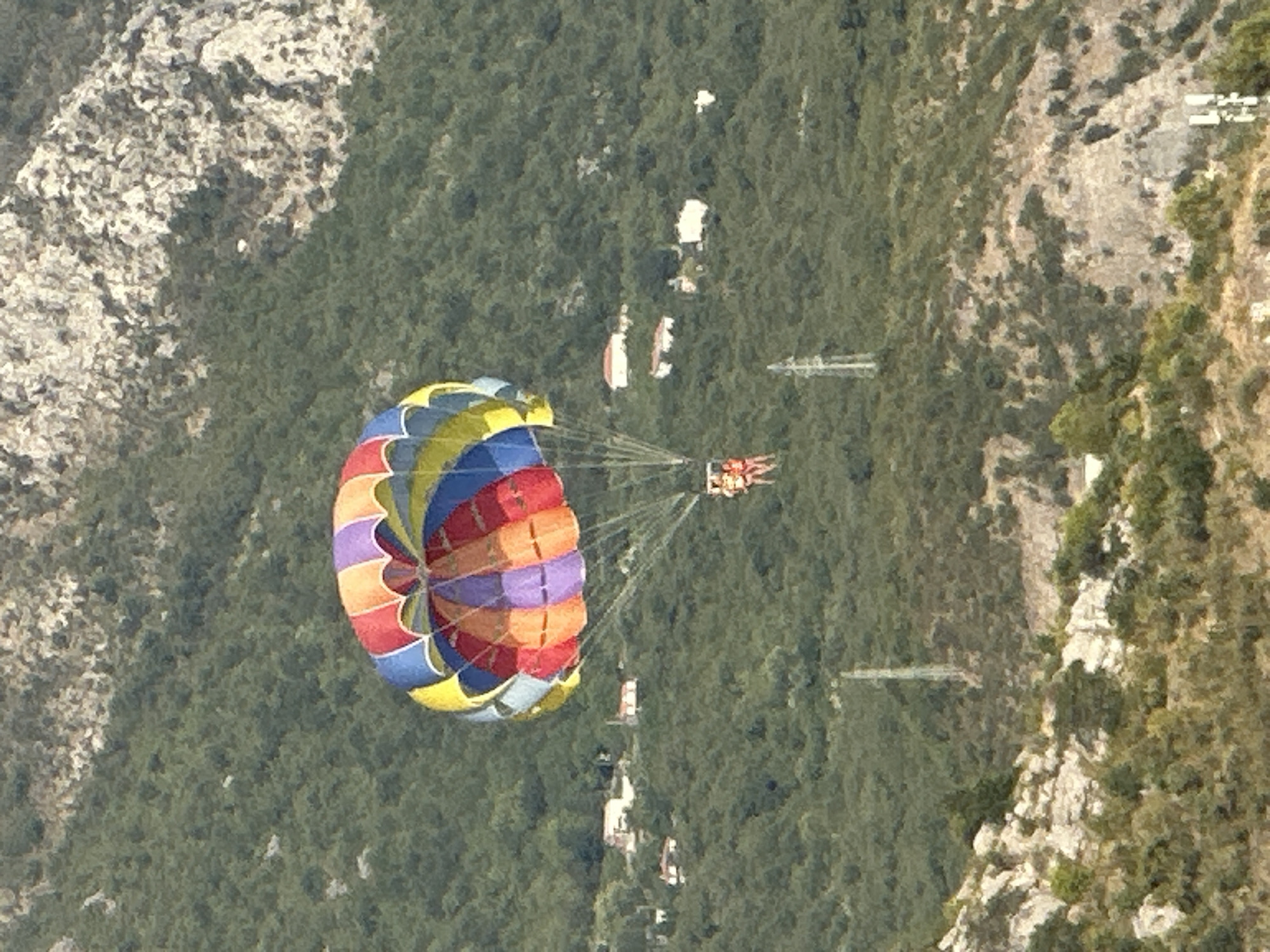 A colorful parasail, with segments of red, orange, yellow, blue, purple, and green, is high in the sky over a lush green hillside dotted with white buildings. Two individuals, wearing orange life vests, are seated in the harness beneath the parasail. The landscape is characterized by dense vegetation covering rolling hills, with exposed rocky outcrops. In the middle ground, several white structures with red roofs are visible, appearing to be houses. Two tall, thin electrical towers stand on the hillside, one prominently in the mid-ground and another further in the background. The lower foreground shows a more arid terrain with sparse vegetation and rocks. The overall scene suggests a recreational activity taking place in a scenic, hilly coastal area.