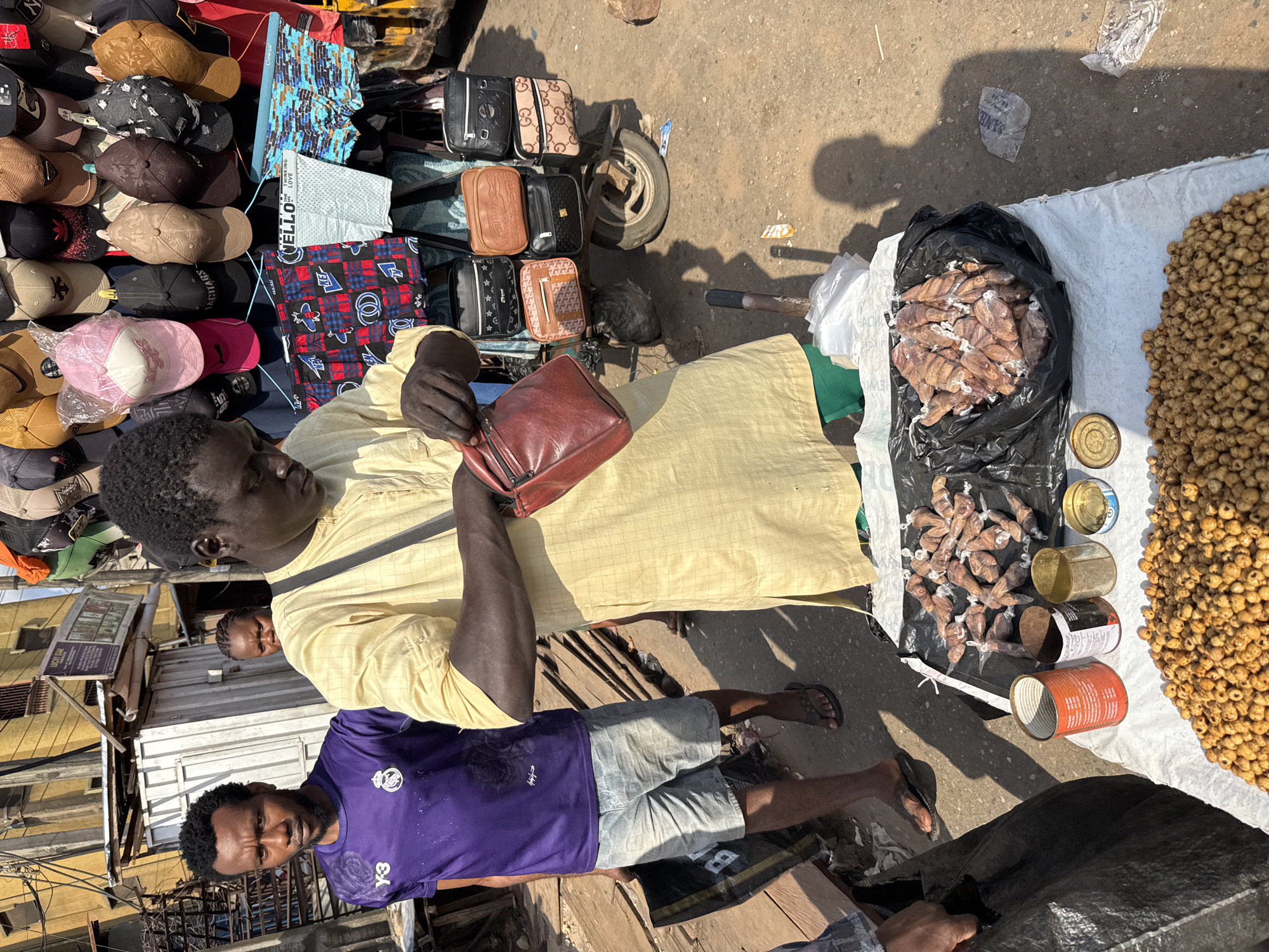 The image captures a street scene in Nkpor, Nigeria. A vendor wearing a yellow shirt is holding a brown purse, presumably selling it alongside other purses displayed on a modified tricycle cart. The cart also holds hats and fabrics. To the right, food items are laid out on a white sheet: bags of unknown contents, several empty cans, and a pile of small, round items. Another person wearing a purple shirt and shorts is standing near the goods on the ground. The environment suggests an outdoor market or commercial area, with other buildings visible in the background.
