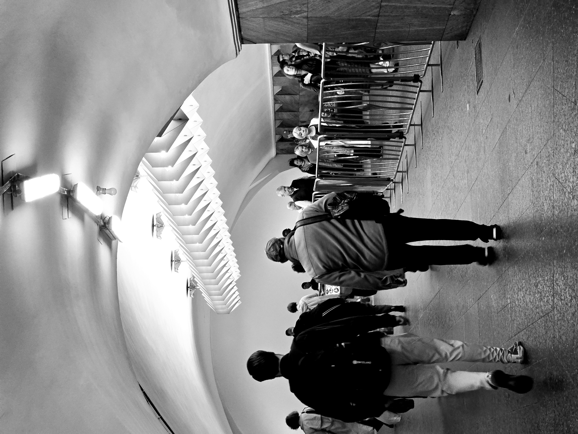 A black and white, angled shot features a line of people walking in a Moscow, Russia, subway station. A curved white ceiling is on the left, adorned with geometric light fixtures. On the right, a metal railing separates the walking area from a crowd of people behind it. The people in the line are dressed in casual clothing and are walking towards the viewer's perspective. The floor is tiled.
