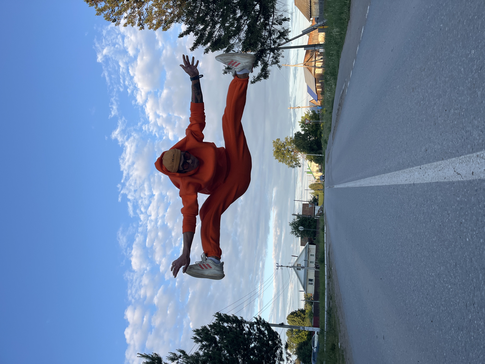 This dynamic outdoor photograph, rotated 90 degrees clockwise from its natural orientation, captures a person in mid-air, performing a high kick or jump against a bright sky. The setting is a road in Chaikovsky, Russia.
The primary subject is a male individual, appearing to defy gravity in a moment of athletic motion. He is dressed in a matching, vibrant orange hooded tracksuit and light-colored sneakers adorned with three red stripes, characteristic of Adidas. A light brown beanie, possibly with "Hollister" text, covers his head, and dark sunglasses obscure his eyes. Tattoos are visible on his forearms, and a watch is worn on his left wrist. His left leg is extended high and straight, while his right leg is bent, and his arms are spread for balance, creating an energetic and acrobatic pose.
The scene is set alongside an asphalt road that features a distinct white dashed line running along its length. The background includes lush green trees, both deciduous and evergreen, as well as utility poles with power lines. In the distance, several houses with peaked roofs are visible, some appearing to be wooden and featuring blue detailing.
The time of day appears to be late afternoon or early evening, characterized by bright, clear blue skies punctuated by scattered white, cumulus clouds. The lighting is strong, casting subtle shadows and highlighting the person's orange attire. The overall mood is one of energetic movement and open-air activity.