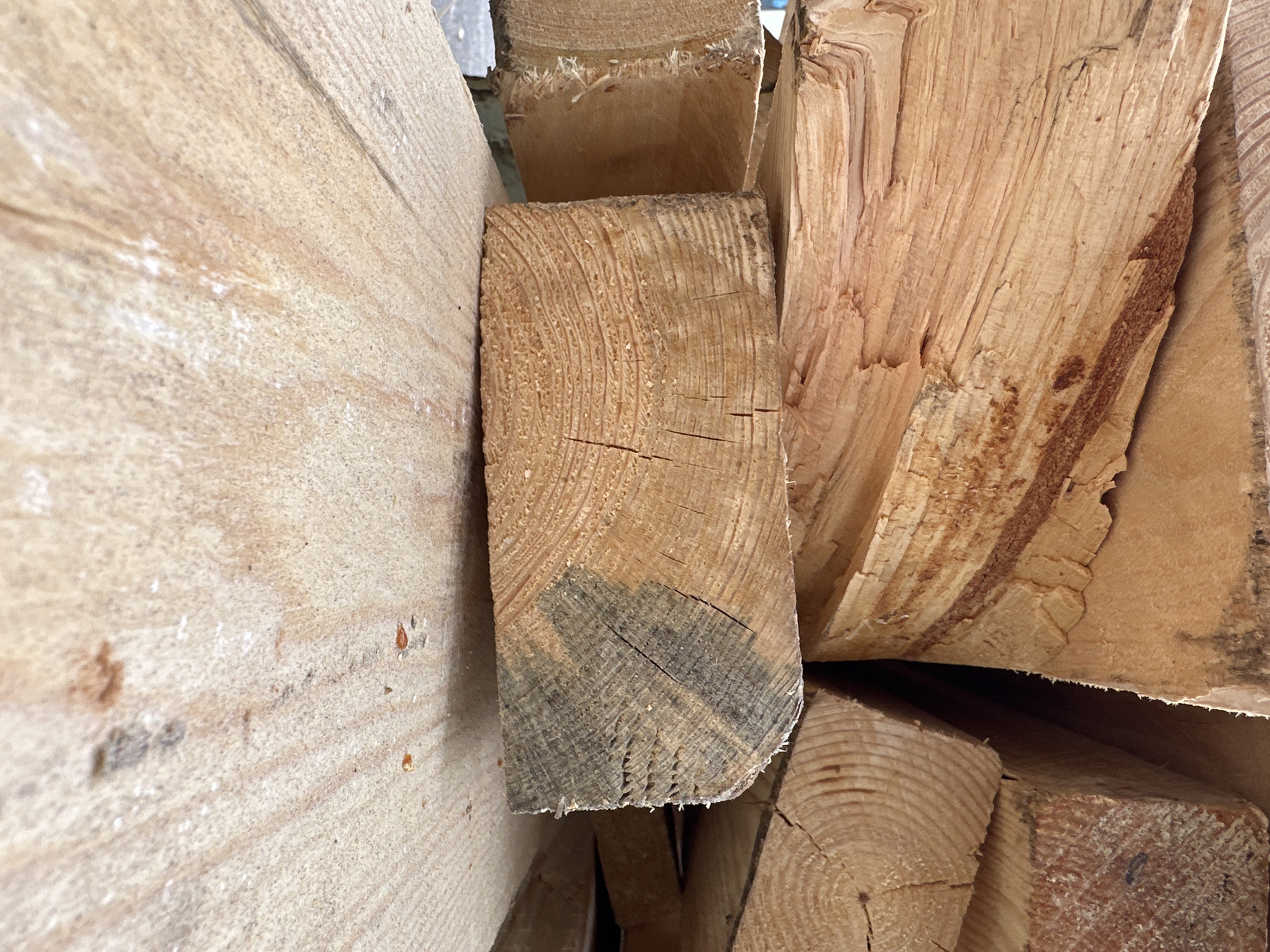 The image shows a close-up view of stacked lumber, likely at a construction site or lumberyard in Wismar, Germany. The wood is light brown and appears rough-sawn, with visible grain patterns and some darker staining on one end of a piece. The lumber is stacked horizontally, with several pieces angled in different directions. The foreground is dominated by a wide, flat piece of lumber with a textured surface. The background is out of focus but suggests more stacked wood. The overall scene is dimly lit, creating shadows and highlighting the textures of the wood. There are no people or animals visible in the image, and no activity is taking place. The environment appears to be outdoors, given the natural light and the presence of what might be a paved or gravel surface in the very bottom left corner.