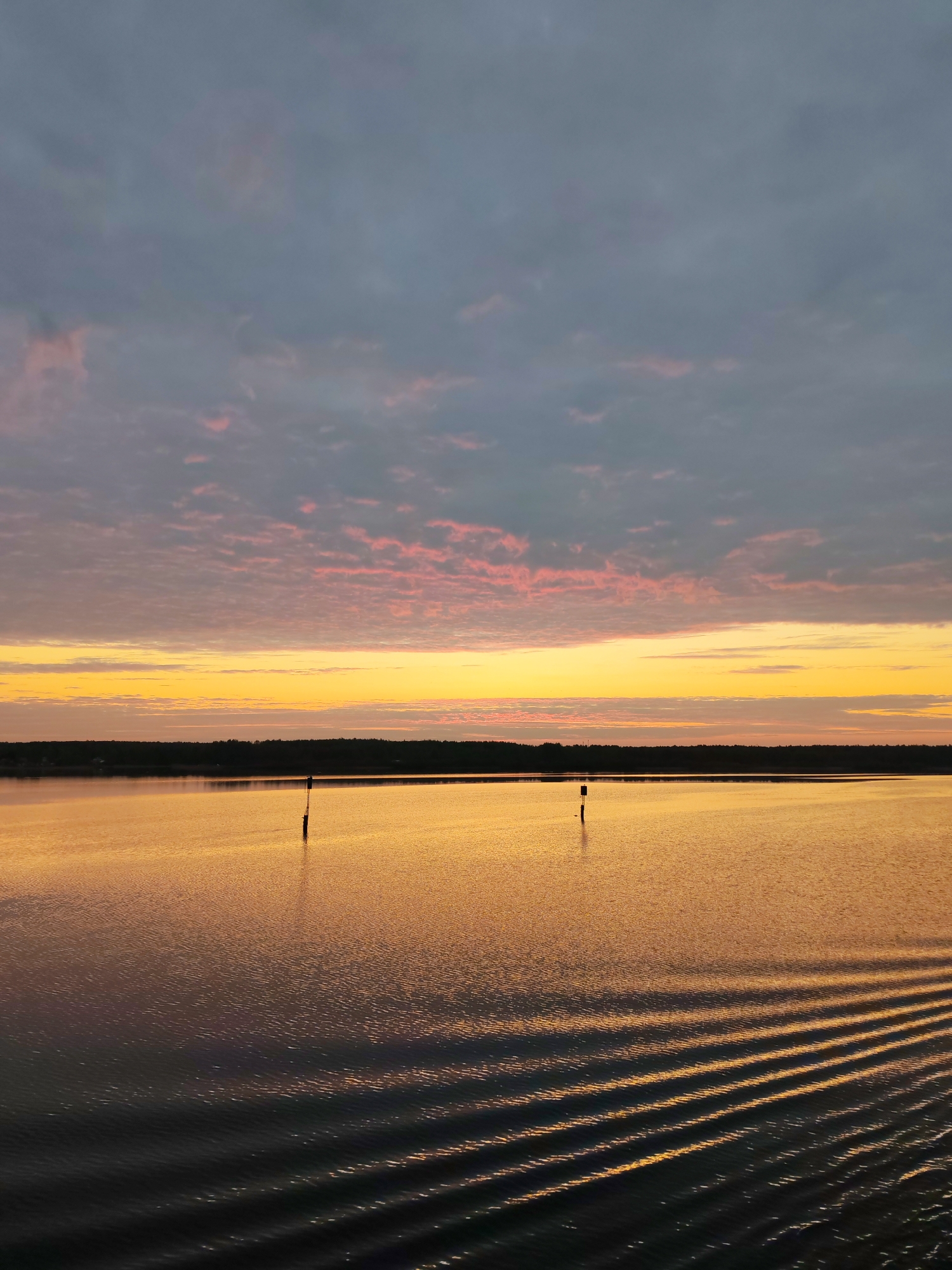 The image shows a body of water at sunset. The sky above the water is filled with clouds, with pink hues near the horizon transitioning to grey at the top. A band of yellow and orange light illuminates the horizon.
Below the horizon, a dark strip of land can be seen. In the water, there are two poles sticking up. The surface of the water is reflective, and lines of ripples are visible in the foreground.
There is no indication of the location of this scene.