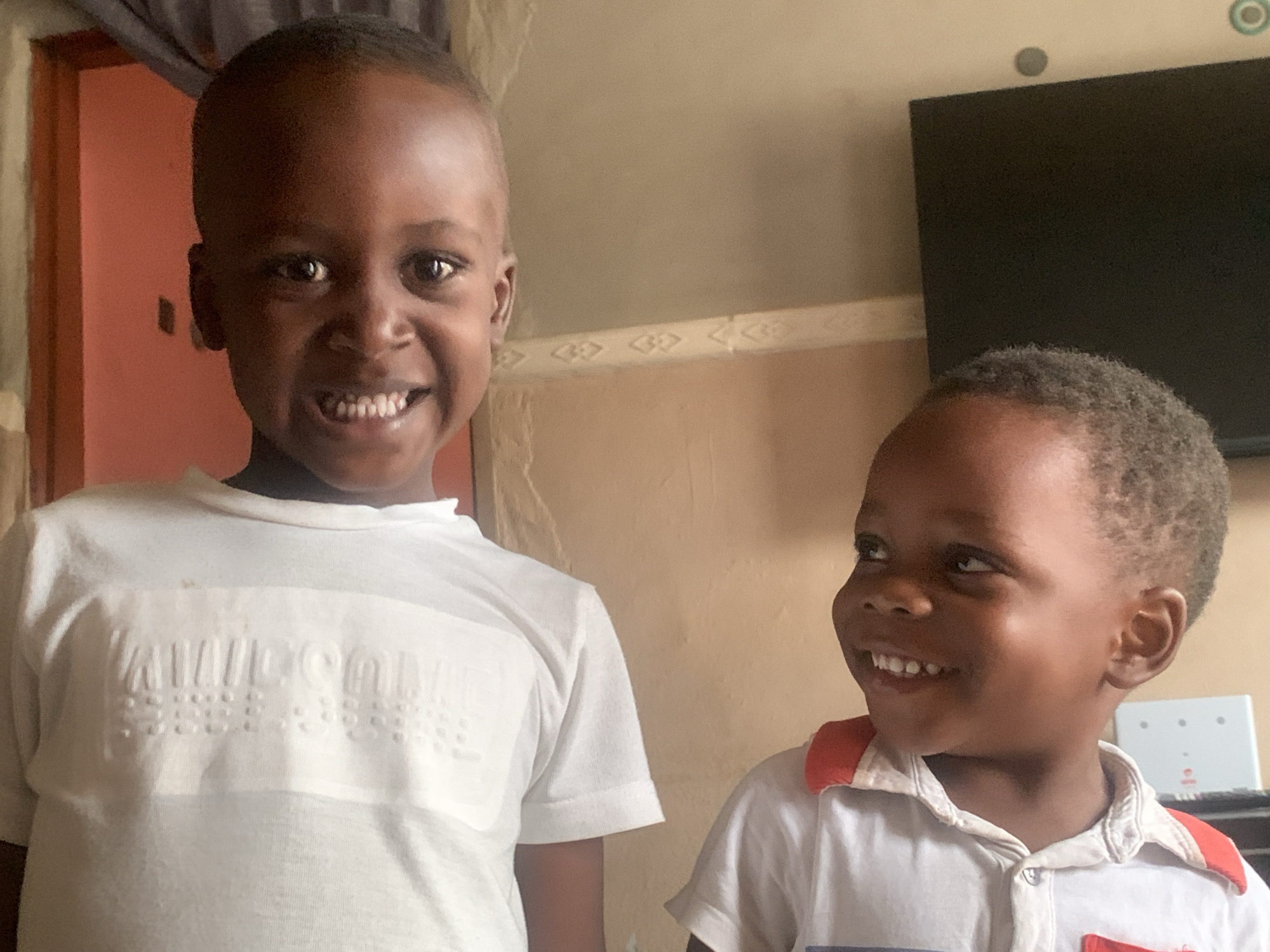 Two young Black boys are shown in a medium close-up, indoors, in what appears to be a residential setting in Sabon Tasha, Nigeria. The boy on the left, older of the two, is looking directly at the camera with a wide smile. He is wearing a white t-shirt with raised text across the chest. His head is shaved. The boy on the right is looking off to his left, also smiling. He has short, curly dark hair and is wearing a white polo shirt with red trim on the collar and a pocket. The background consists of a plain, light brown wall. To the left of the frame, a portion of a red-painted door or wall is visible. To the right, a portion of a black television screen is visible above the younger boy's head. The overall lighting suggests indoor illumination.