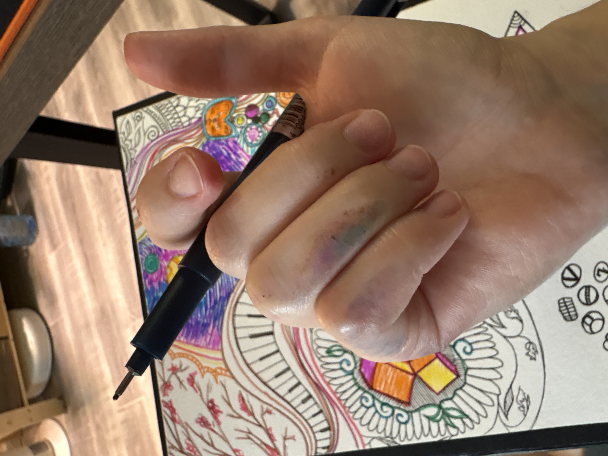 A close-up shot displays a hand holding a pen over a drawing. The drawing features intricate patterns and designs, including elements resembling piano keys, geometric shapes, and floral motifs. The pen is black with a metallic tip. The hand appears to have some bruising on the knuckles. The background is blurred, showing parts of a desk or table and other drawing materials. There is no information available to determine the location as Saint Petersburg, Russia.