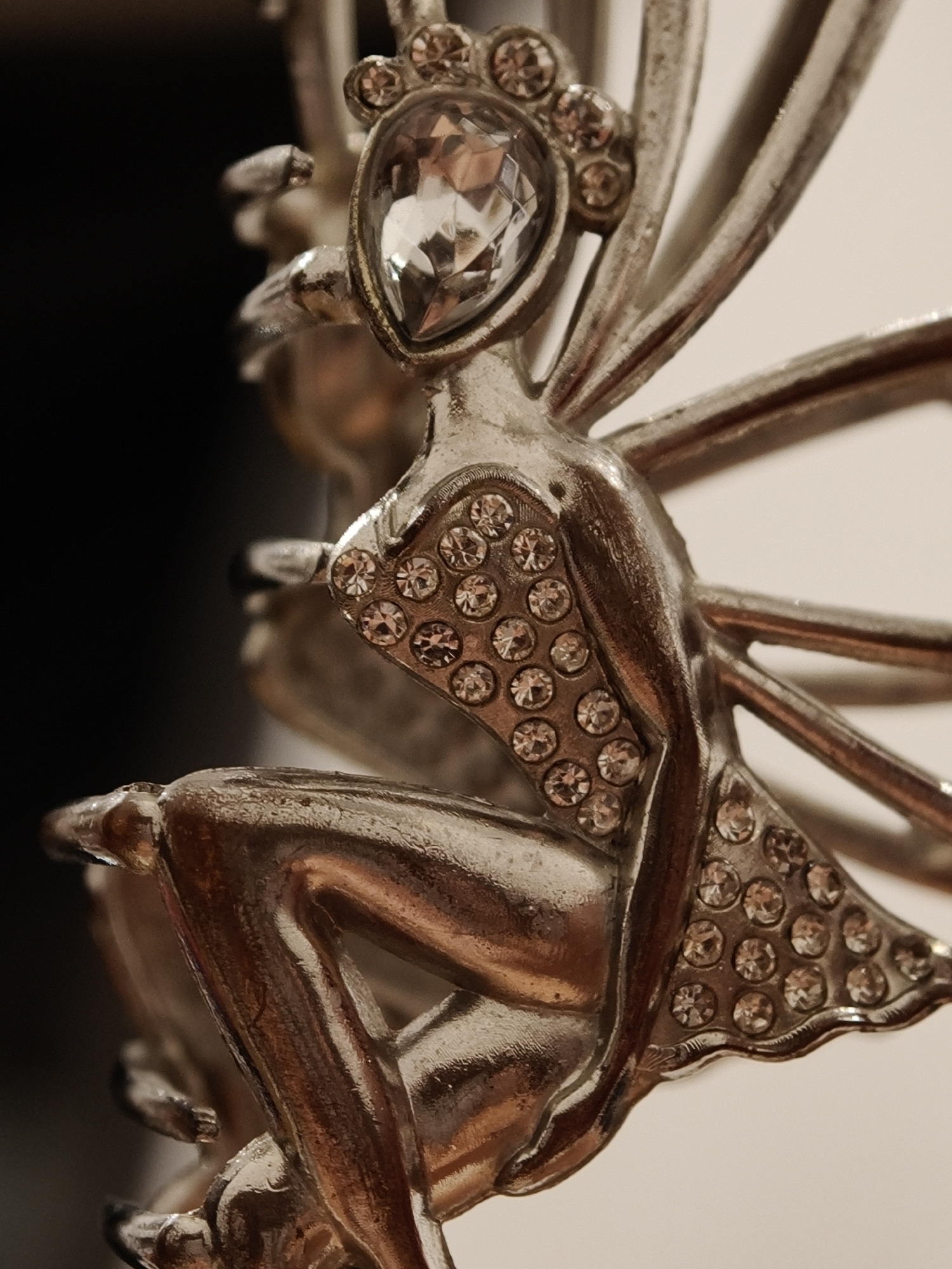 The image shows a close-up, macro photograph of a silver-toned decorative object resembling a fairy. The fairy is intricately detailed, with a prominent faceted gem at the center of its head, surrounded by smaller sparkling gems. The fairy's body and wings are also adorned with numerous small, clear gems arranged in circular patterns. The fairy is depicted in a seated position, with its legs tucked beneath it. The object appears to be made of metal and is highly reflective, with highlights and shadows emphasizing its metallic texture. The background is a soft blur of dark and light colors, suggesting an indoor setting with controlled lighting. There is no indication of the object's location in the city of Đà Nẵng, Vietnam; it is a close-up of a decorative item.