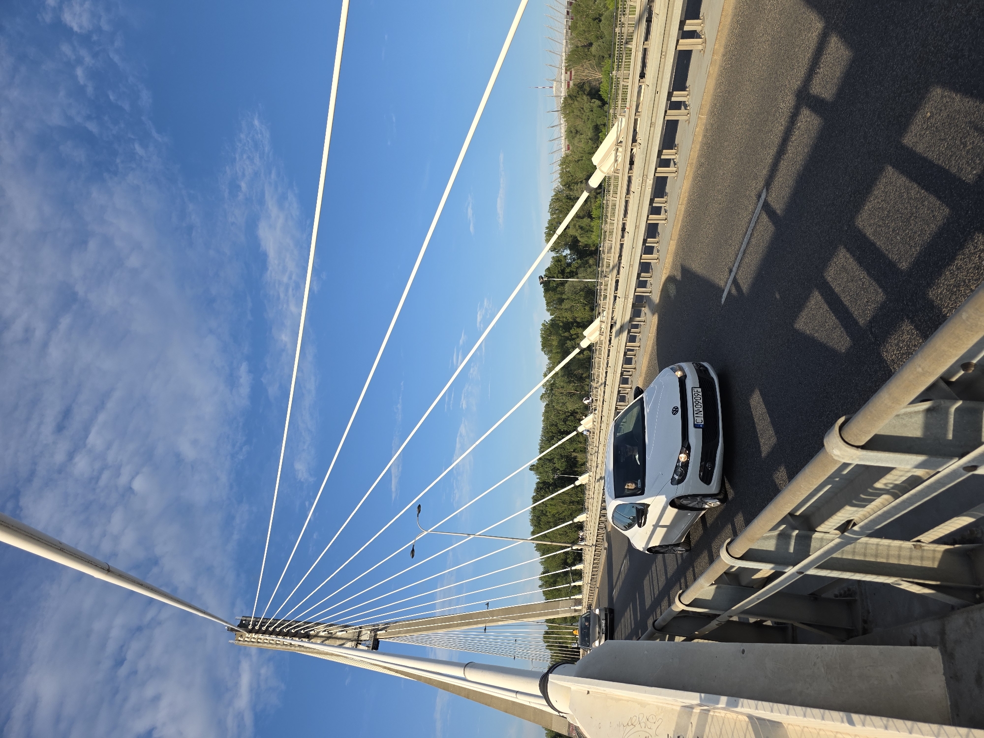 The image shows a view of the Świętokrzyski Bridge in Warsaw, Poland, on a sunny day. The bridge is a cable-stayed bridge with a prominent pylon and numerous white cables extending to the deck. The sky is blue with scattered white clouds.
In the foreground, a white Volkswagen Scirocco is driving on the bridge. The car has its headlights on and a license plate that reads "CIN-0909F". The driver is visible behind the wheel. The road is asphalt and appears to be in good condition.
In the background, on the right side of the image, a stadium structure with a distinctive roof can be seen. Trees and vegetation line the banks of the river or body of water that the bridge spans. Another car, a silver sedan, is visible further ahead on the bridge. There are also some distant figures walking on the sidewalk of the bridge. The overall impression is that of a busy urban environment with modern infrastructure.